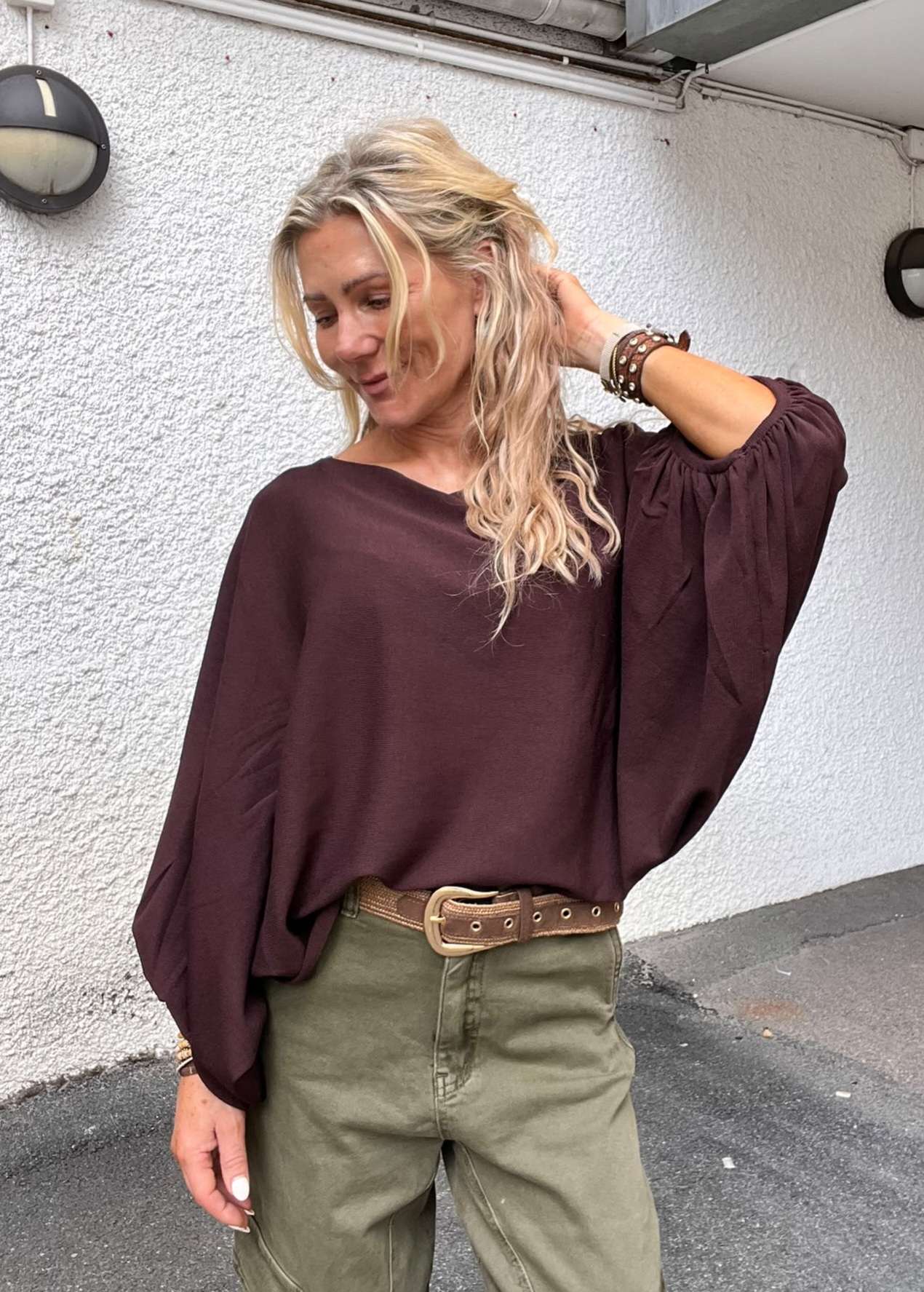 Blond kvinna står avslappnat och rör vid håret med ena handen, medan hon tittar ner och ler svagt. Hon bär vinröd, lös blus med vida ärmar, olivgröna byxor och brunt bälte, framför vit yttervägg.