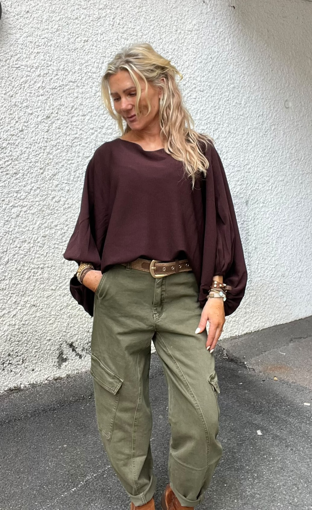 Blond kvinna står avslappnat mot en vit, grovputsad vägg och tittar nedåt. Hon har vinröd, vid blus med ballongärmar, olivgröna cargobyxor med brunt skärp, armband och bruna skor på asfalt.