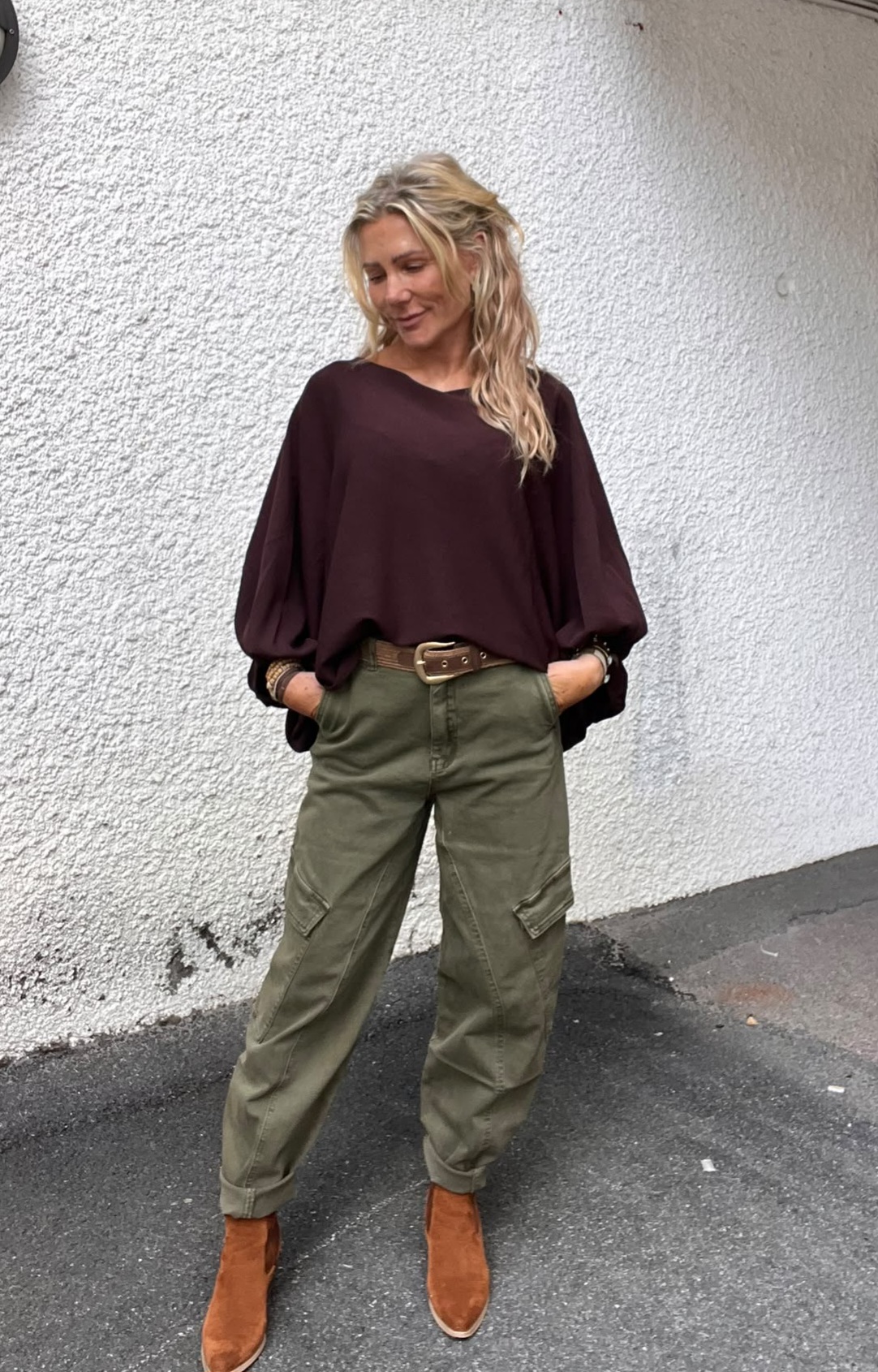 Blond kvinna står avslappnat med händerna i fickorna och tittar nedåt med ett svagt leende. Hon bär vinröd, luftig blus, gröna cargobyxor med bälte och bruna mockakängor mot en vitputsad vägg.
