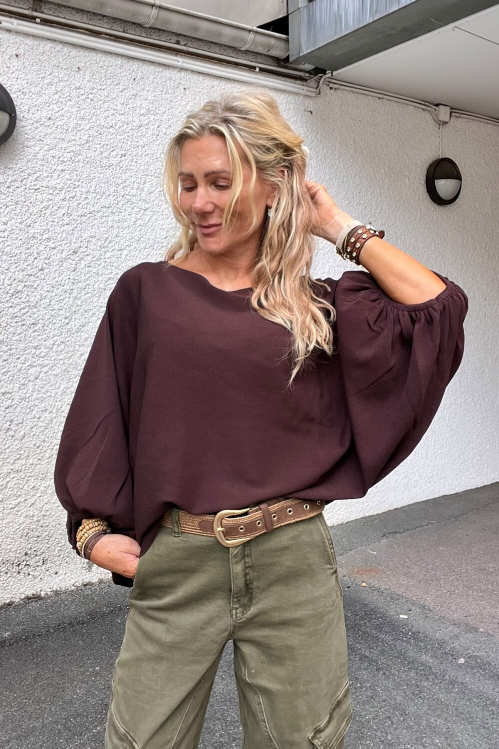 Blond kvinna står utomhus vid en vit husvägg och lutar sig avslappnat, med ena handen i fickan och den andra i håret. Hon bär vinröd blus, gröna byxor, bredt bälte och armband.