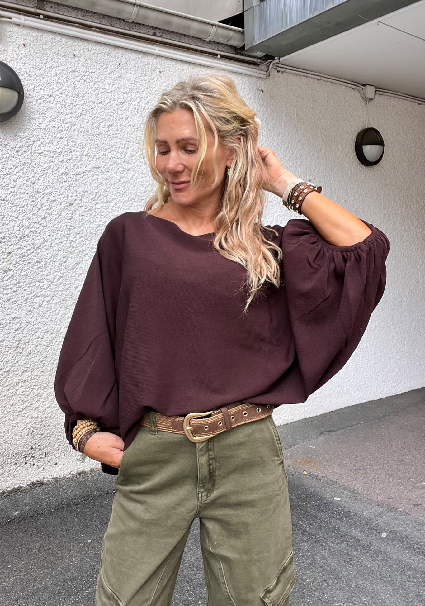 Blond kvinna står utomhus vid en vit husvägg och lutar sig avslappnat, med ena handen i fickan och den andra i håret. Hon bär vinröd blus, gröna byxor, bredt bälte och armband.