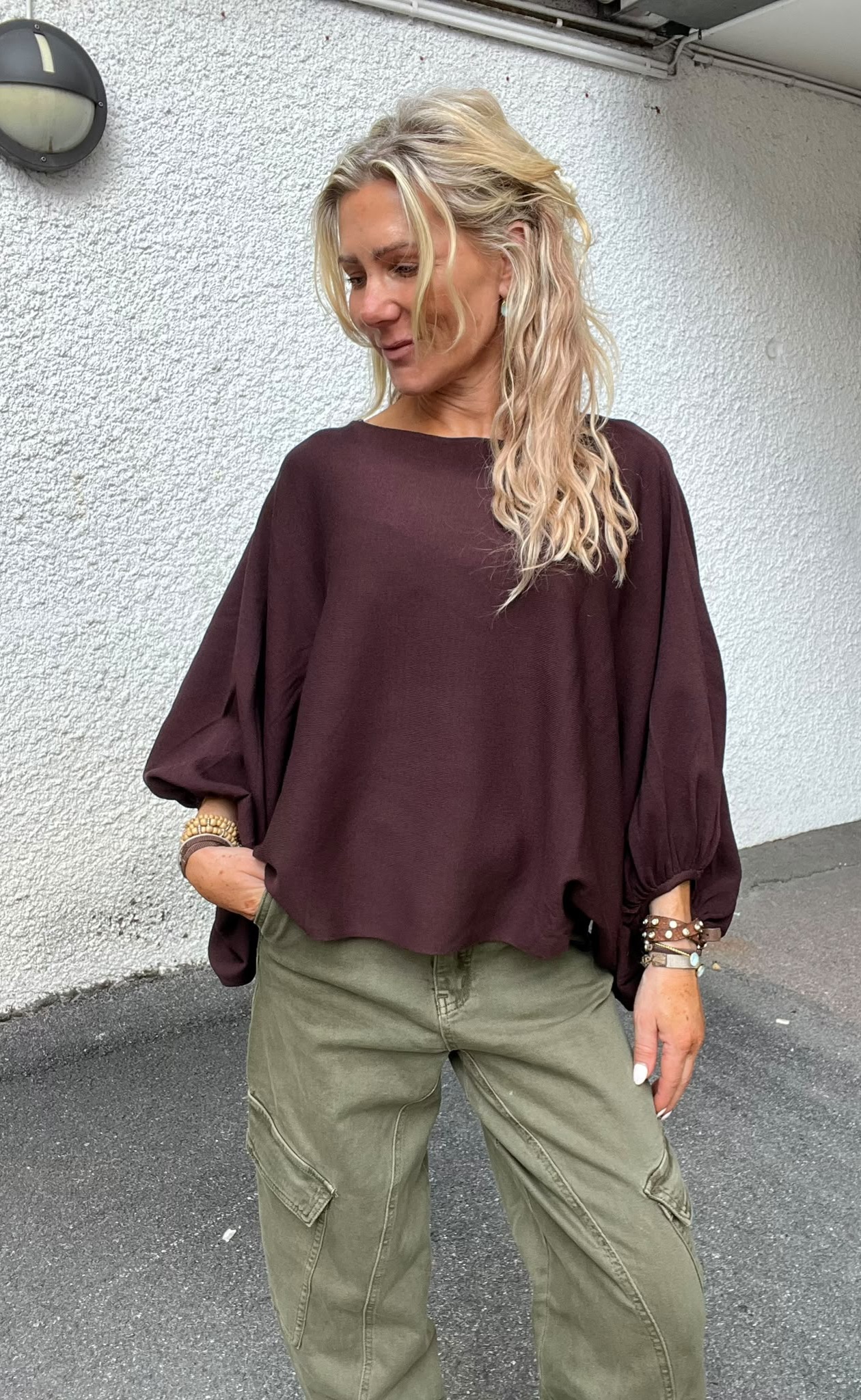 Blond kvinna står utomhus vid en vit, grovputsad vägg och tittar nedåt med ett svagt leende. Hon har en vinröd, lös topp med puffärmar och olivgröna cargobyxor samt flera armband.