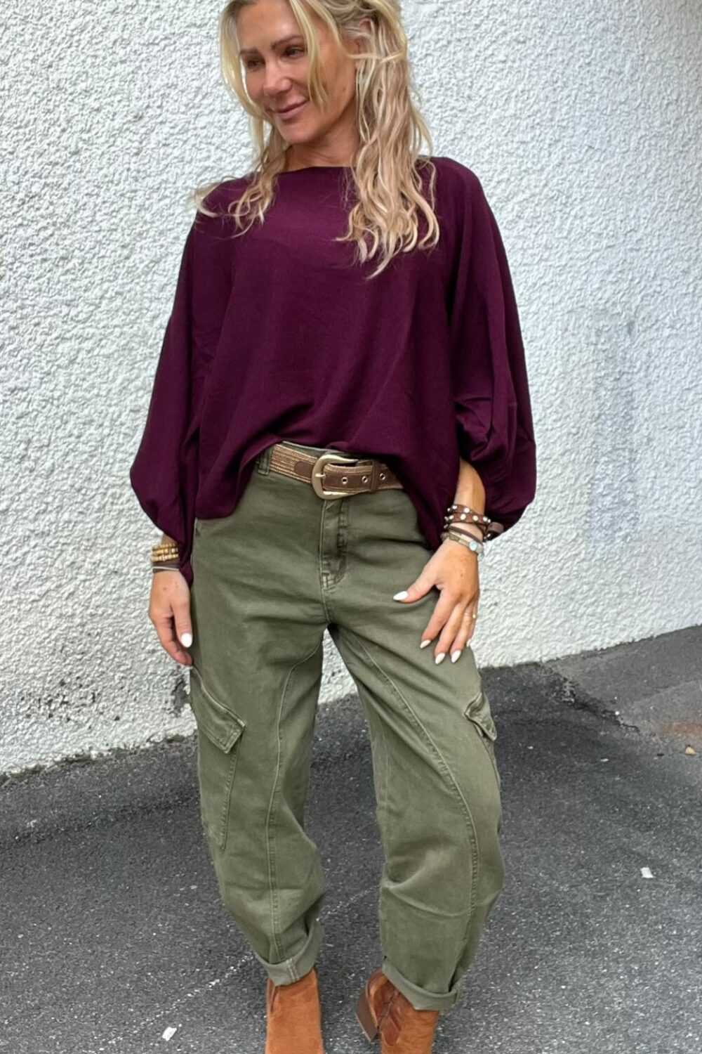 Blond kvinna poserar utomhus mot en vit, grovputsad vägg och tittar åt sidan med ett leende. Hon bär vinröd långärmad blus, olivgröna cargobyxor med bälte, bruna ankelboots och flera armband.