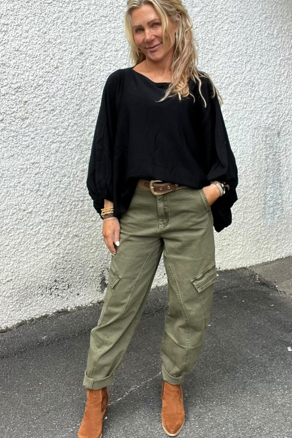 Blond kvinna står avslappnat mot en ljus, grovputsad vägg och ler mot kameran, med ena handen i fickan. Hon bär svart blus, gröna cargobyxor med brunt bälte och bruna ankelboots på asfalt.