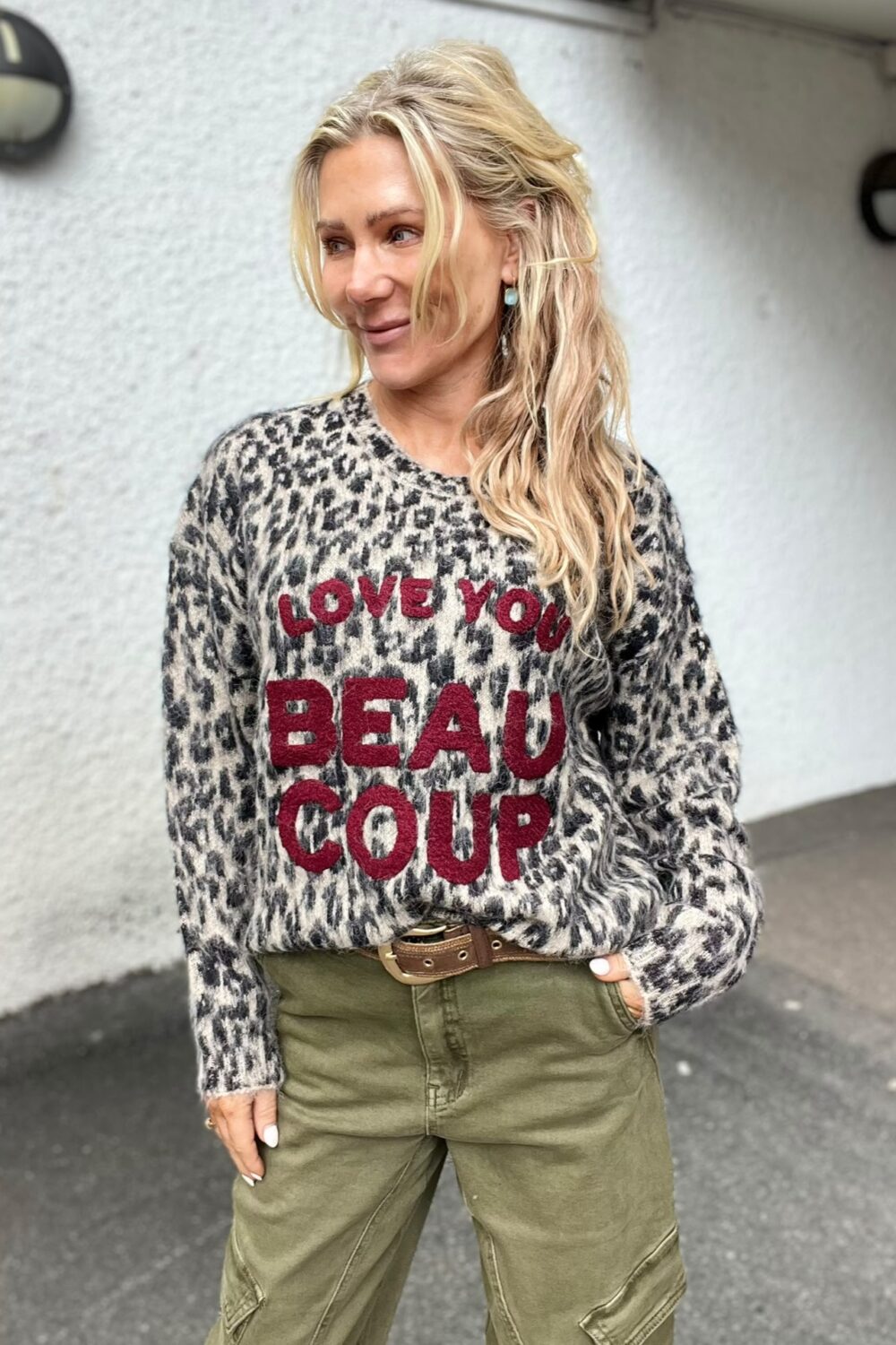 Blond kvinna står utomhus och ler åt sidan med ena handen i fickan. Hon bär leopardmönstrad stickad tröja med röd texten ”LOVE YOU BEAU COUP” och olivgröna byxor mot ljus vägg.