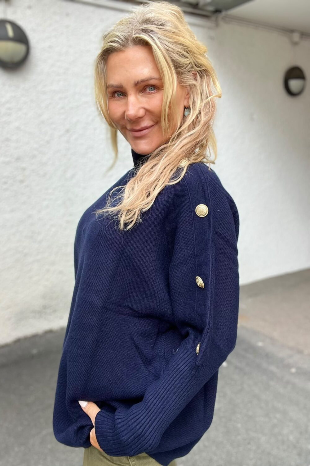 Blond kvinna i marinblå stickad tröja med guldfärgade knappar och örhängen står utomhus vid en vit vägg, vänder sig mot kameran och ler. Bilden är ett halvkroppsporträtt med lampor på väggen i bakgrunden.