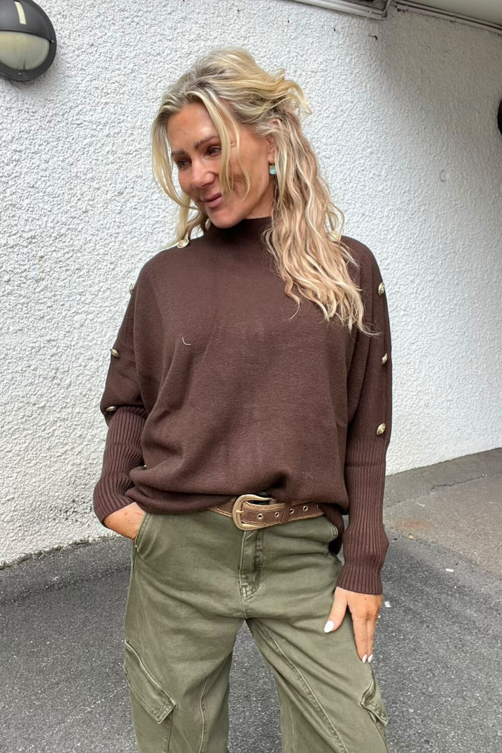 Blond kvinna står avslappnat med ena handen i fickan och ler svagt, klädd i brun stickad tröja med guldfärgade knappar, gröna cargobyxor och brett bälte, mot en ljus, grovputsad vägg utomhus.