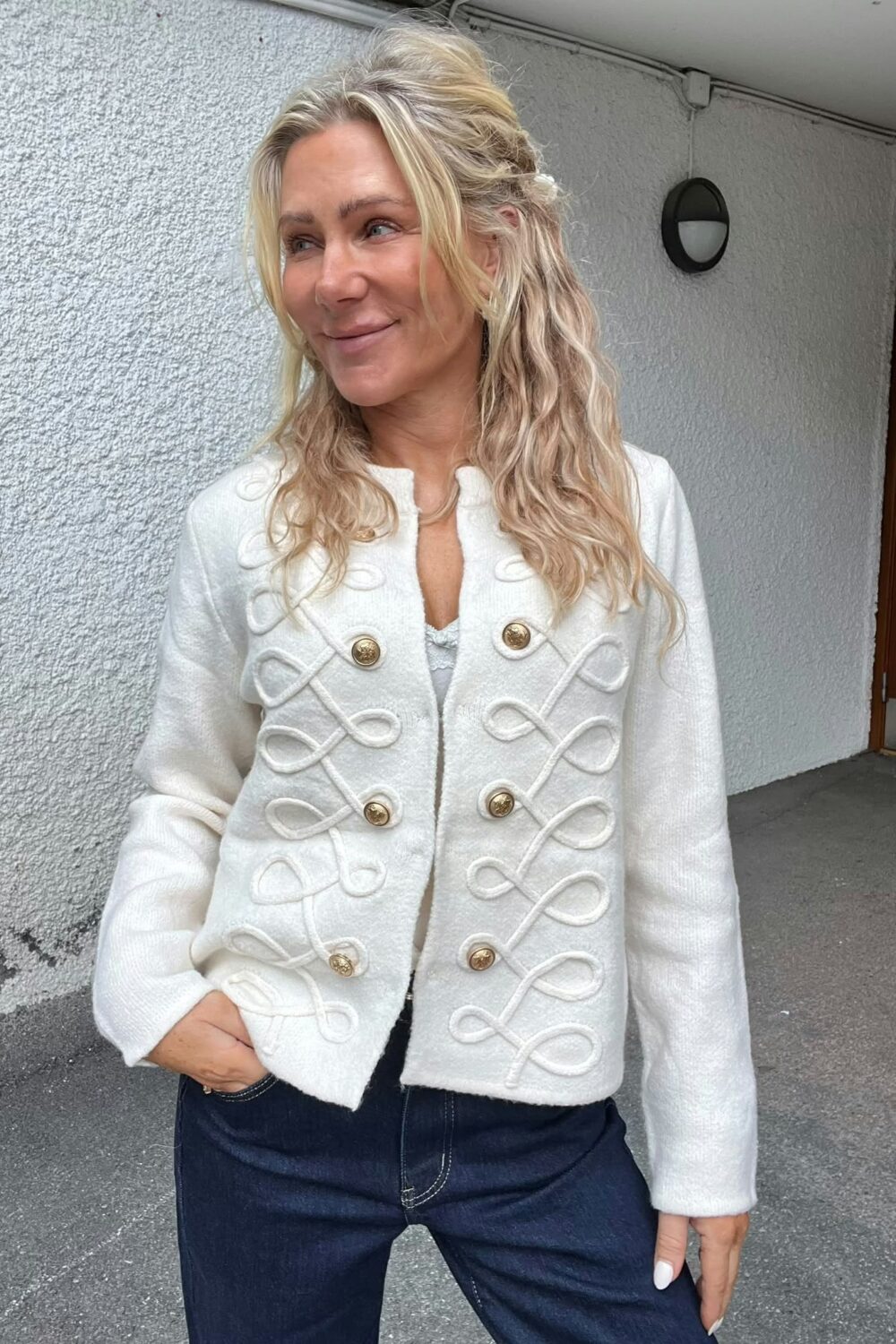 Blond kvinna står utomhus vid en ljus, grovputsad vägg och ler medan hon tittar åt sidan. Hon bär en vit, kort jacka med dekorativa snördetaljer och guldknappar samt mörkblå jeans.