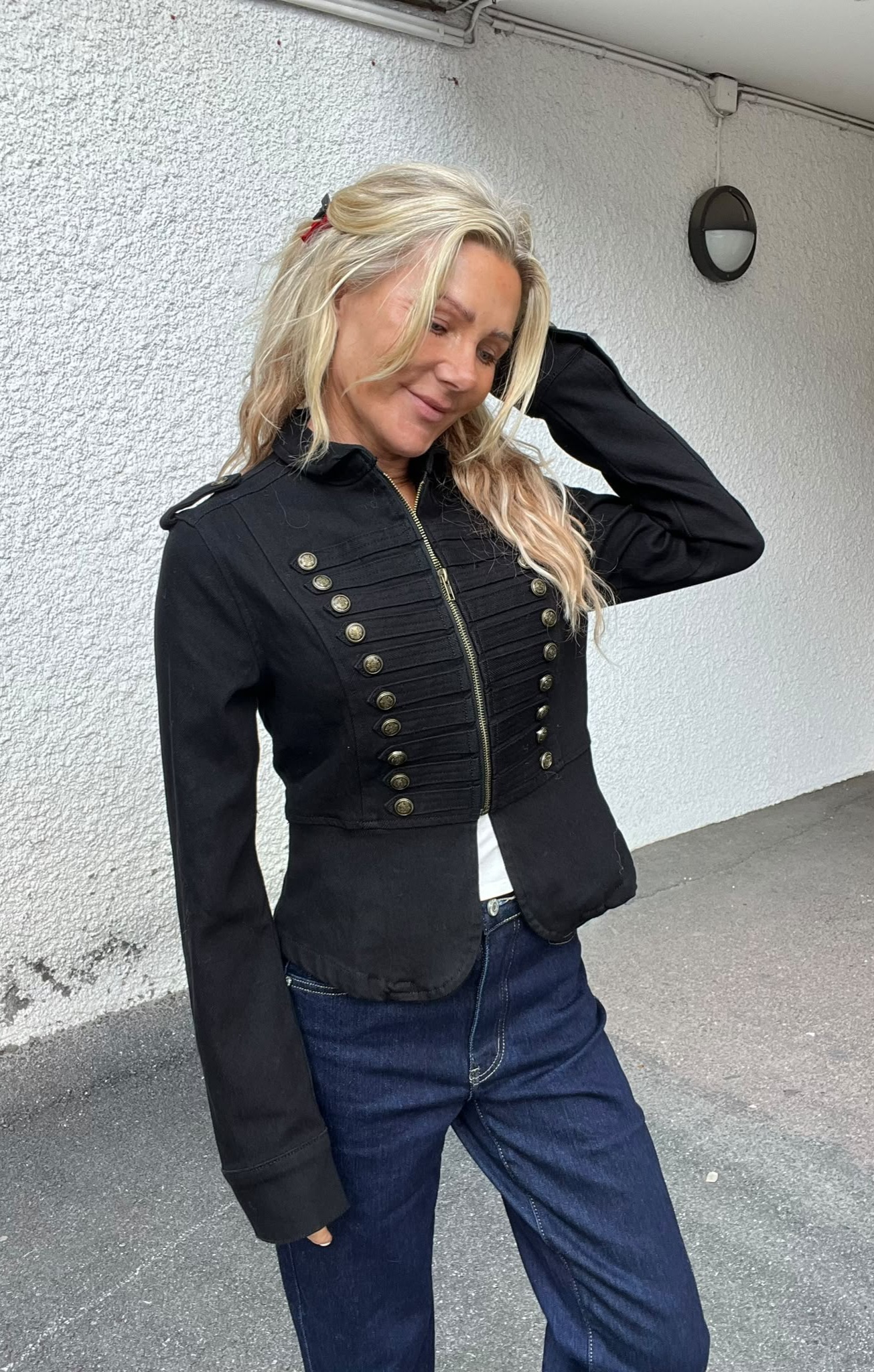 Blond kvinna poserar lutad mot en vit, grovputsad vägg och håller ena handen bakom huvudet. Hon bär en svart, figursydd jacka med dragkedja och dekorativa knappar samt blå jeans; utomhus vid en vägglampa.