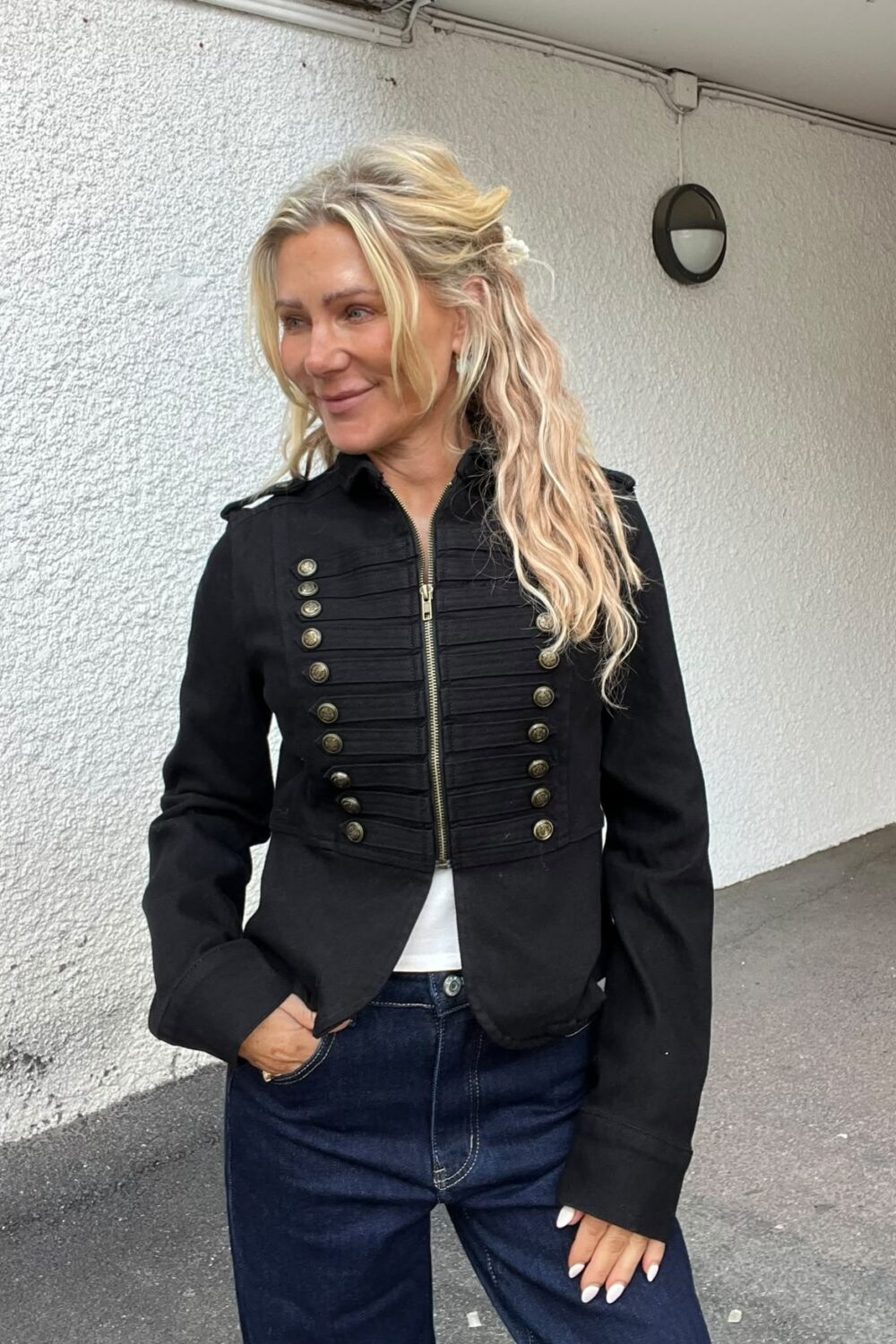 Blond kvinna står utomhus vid en vit putsad vägg och ler medan hon tittar åt vänster. Hon har handen i jeansfickan och bär en svart jacka med guldfärgade knappar och dragkedja.