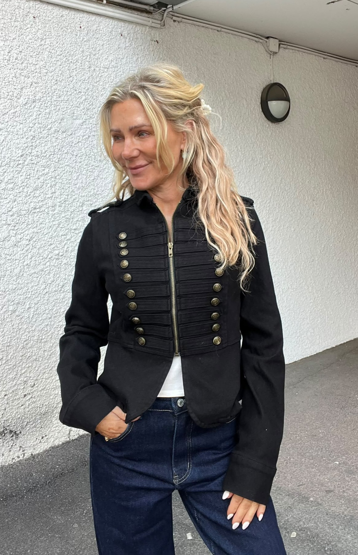 Blond kvinna står utomhus vid en vit putsad vägg och ler medan hon tittar åt vänster. Hon har handen i jeansfickan och bär en svart jacka med guldfärgade knappar och dragkedja.