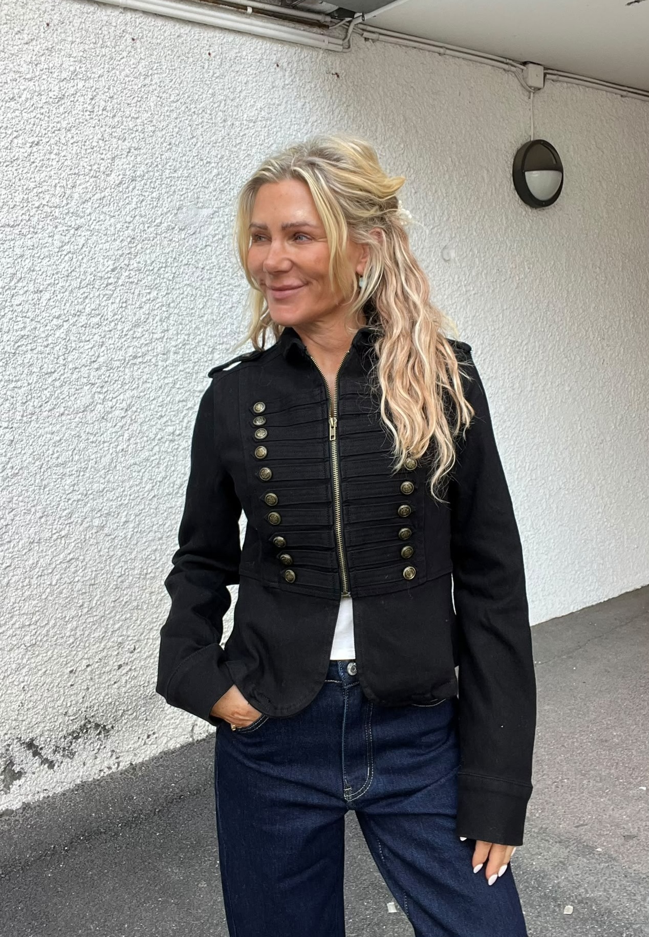 Kvinna med långt blont hår står och ler, med ena handen i fickan. Hon bär en svart, militärinspirerad jacka med guldfärgade knappar och dragkedja samt blå jeans, framför en vitputsad vägg utomhus.