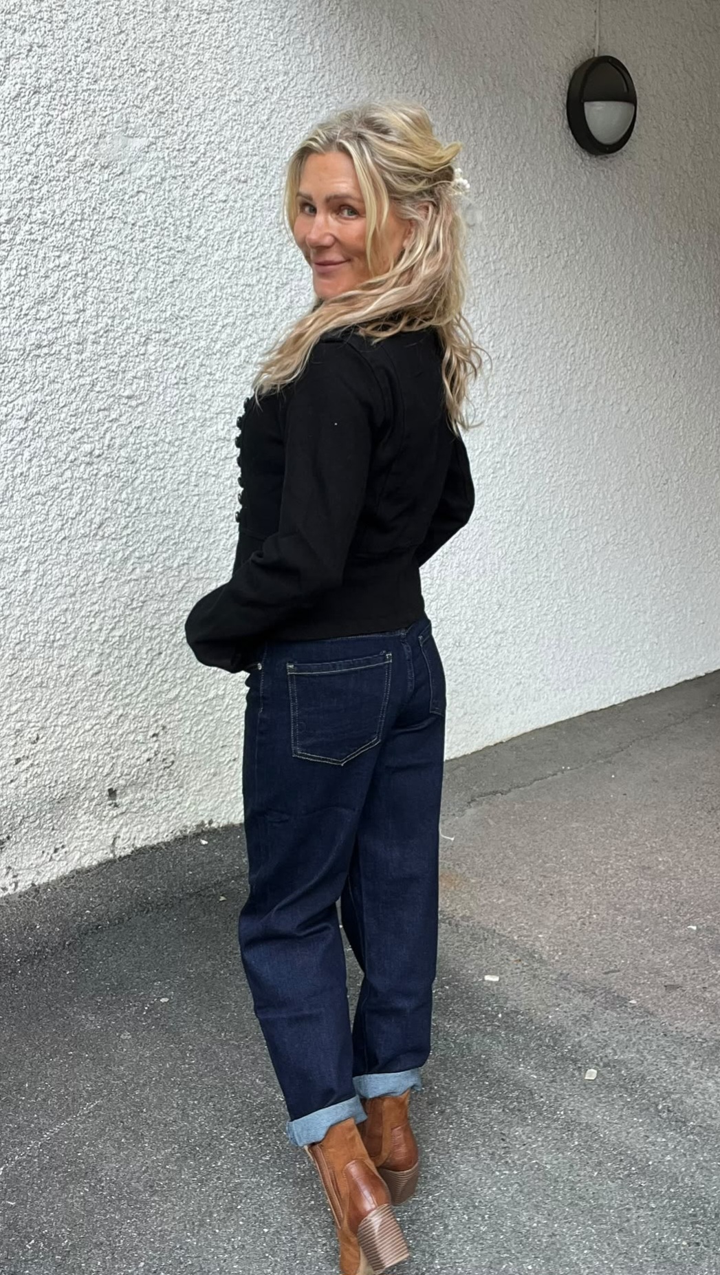 Blond kvinna står i profil och vänder sig om med ett leende mot kameran, klädd i svart blus, mörkblå uppvikta jeans och bruna boots. Hon står utomhus vid en vit, grovputsad vägg med vägglampa.