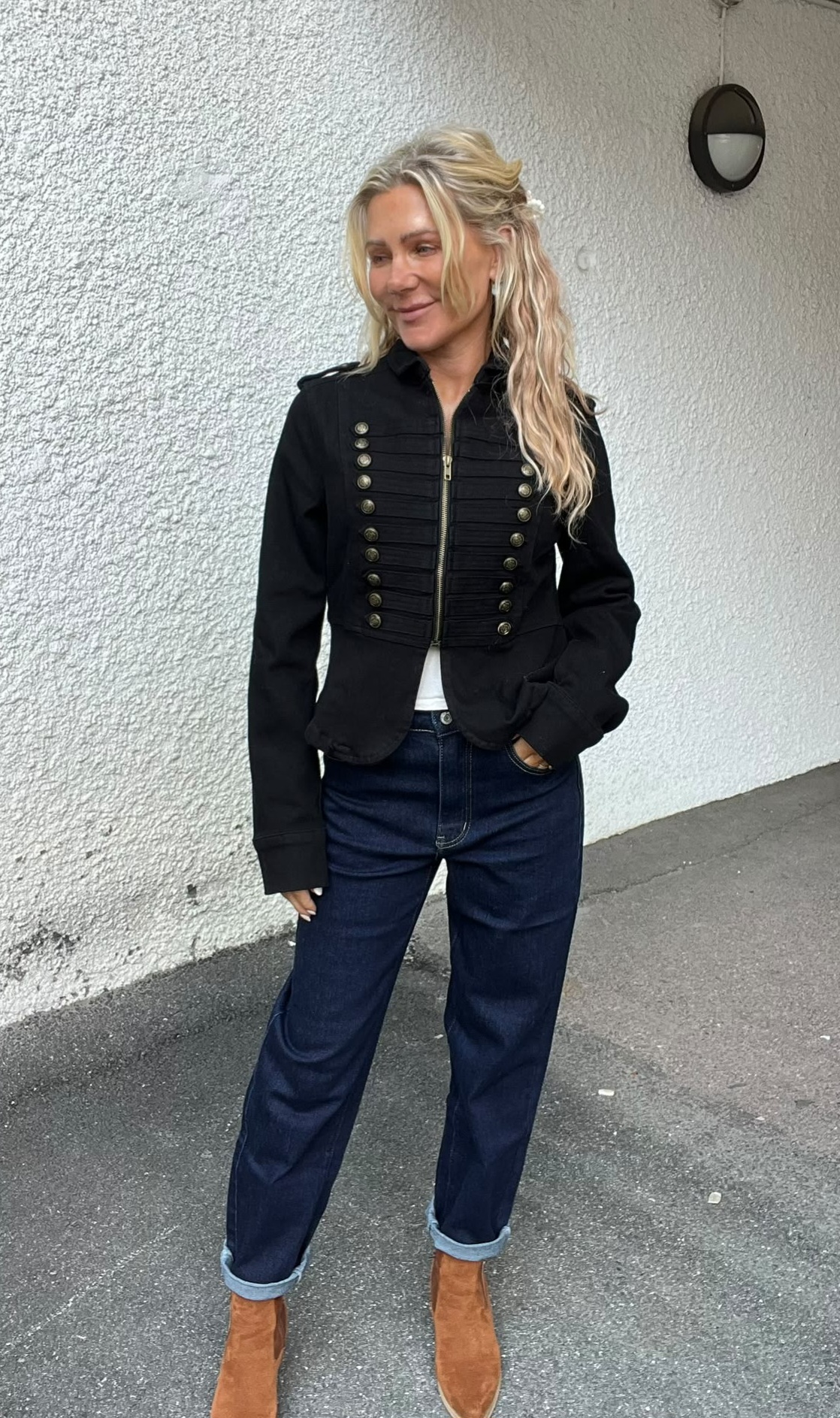 Blond kvinna står avslappnat med ena handen i jeansfickan och ler åt sidan, klädd i svart militärinspirerad jacka med knappar, mörkblå uppvikta jeans och bruna mockaboots, mot en vit putsad vägg utomhus.