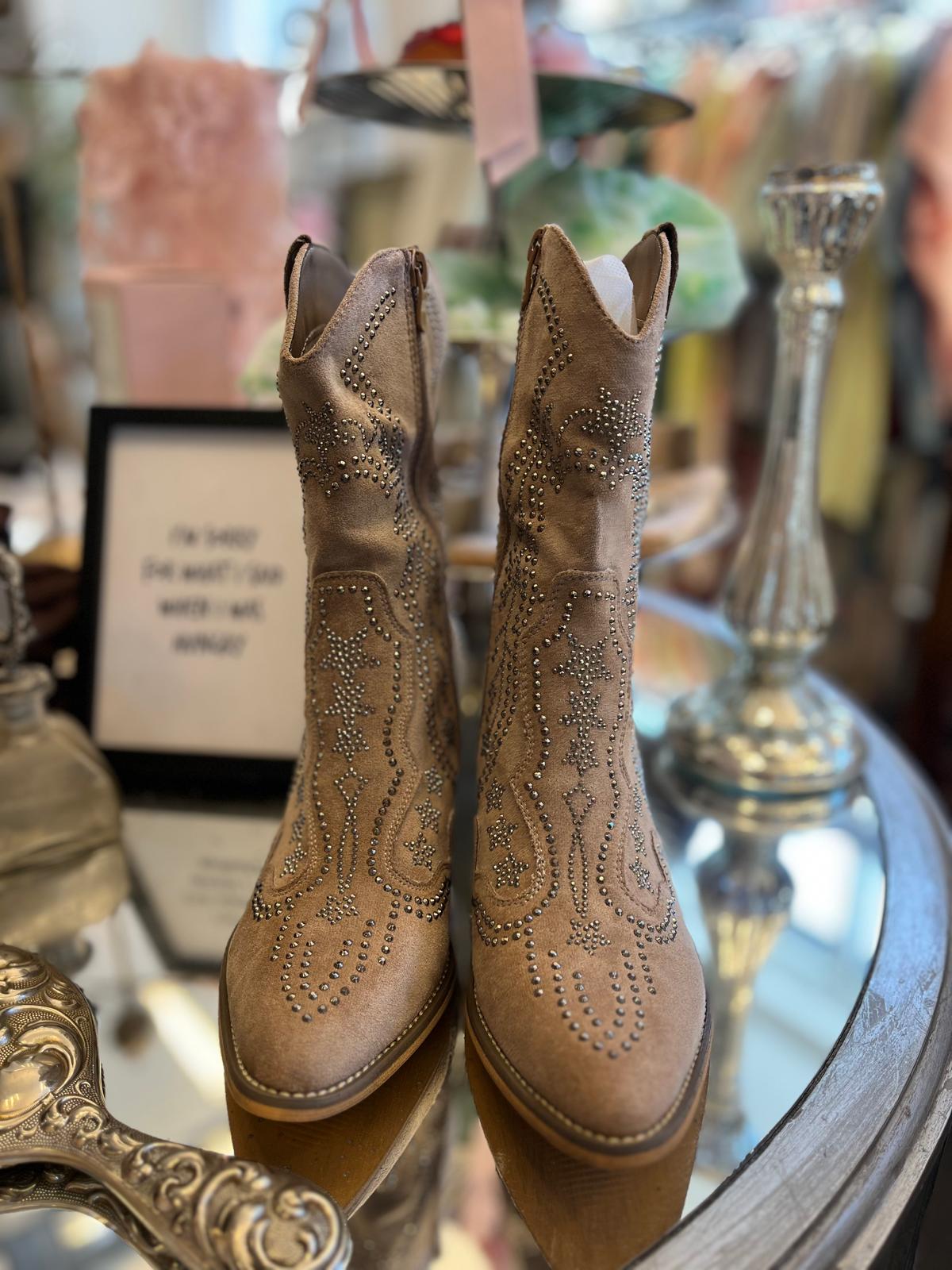 Ett par beige mockaboots med västerländskt mönster i silvernitar står uppställda på ett runt glasbord. I bakgrunden syns en suddig skylt med text och en silverfärgad ljusstake i en butiksmiljö.