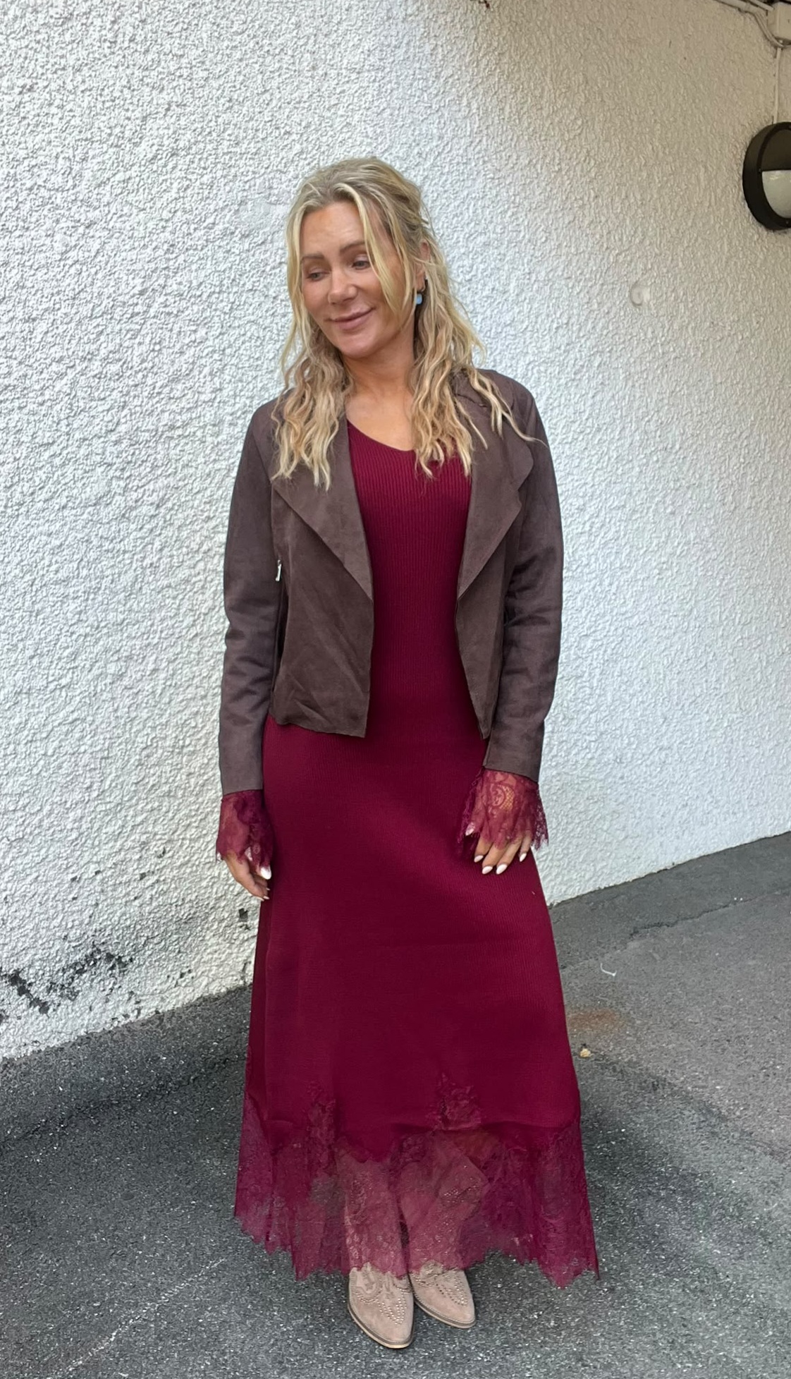 Kvinna med långt blont hår står och ler lätt, vänd åt sidan, mot en vit putsad vägg utomhus. Hon bär vinröd maxiklänning med spetsdetaljer och en brun jacka, samt ljusa boots.