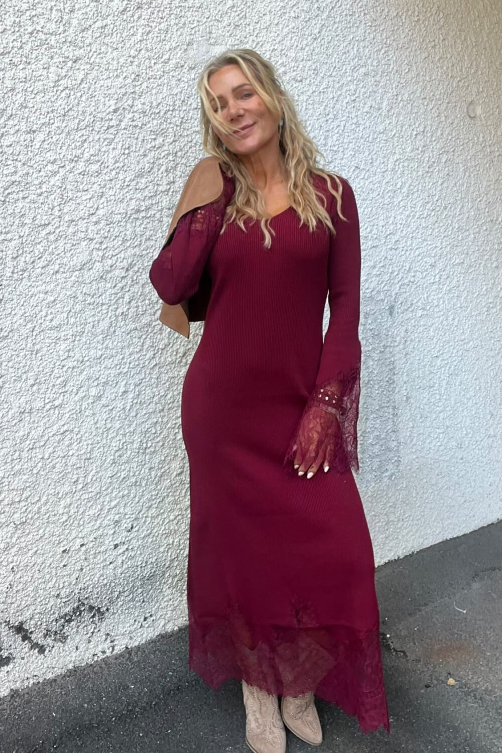 Blond kvinna står lutad mot en vit, grovputsad vägg och ler svagt. Hon bär en vinröd lång klänning med spetsdetaljer vid ärmar och fåll, beige boots och har en brun jacka över axeln.
