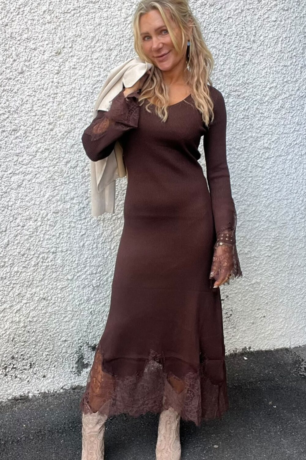 Blond kvinna står och ler mot kameran med en ljus kavaj slängd över ena axeln, klädd i en lång brun klänning med spetsdetaljer och ljusa boots, framför en vit, grovputsad vägg.