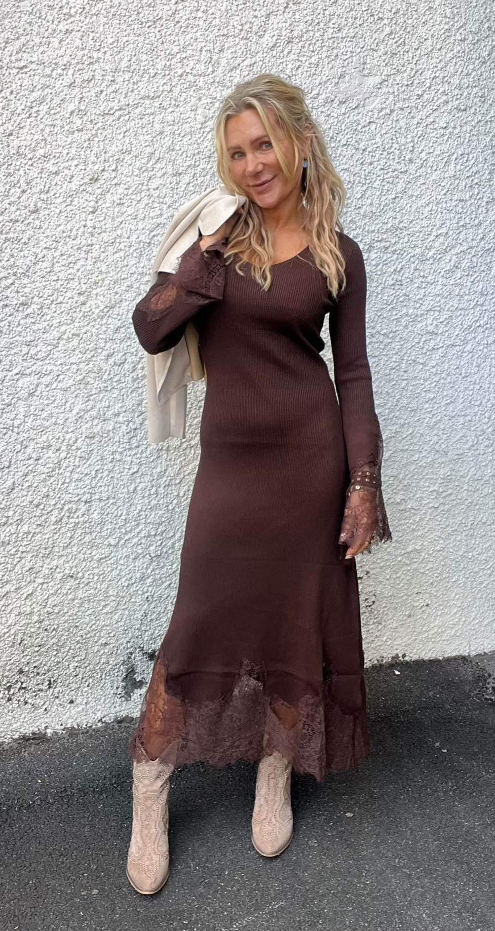 Blond kvinna står och ler mot kameran med en ljus kavaj slängd över ena axeln, klädd i en lång brun klänning med spetsdetaljer och ljusa boots, framför en vit, grovputsad vägg.