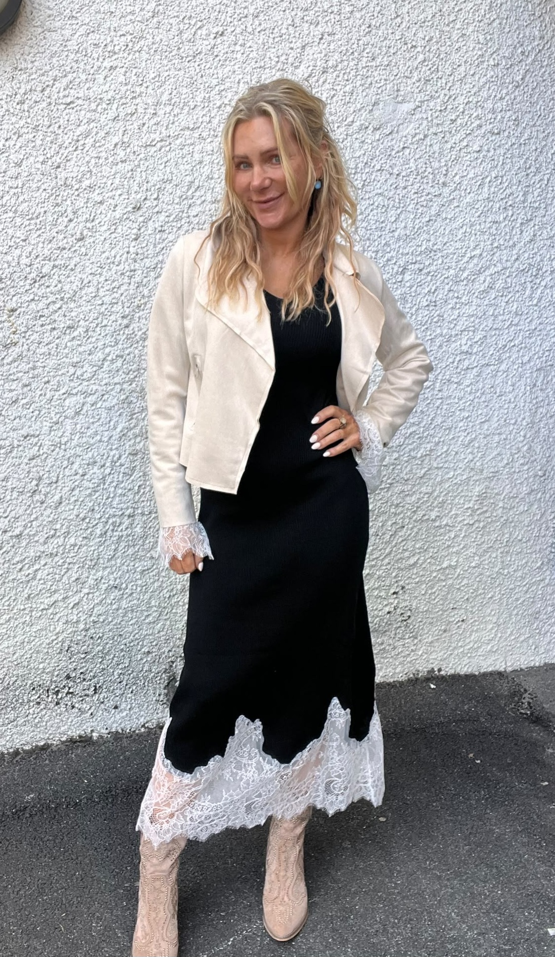 Kvinna med långt blont hår poserar leende med handen på höften framför en vit putsad vägg. Hon bär svart klänning med vit spetskant, ljus jacka och beige boots på asfalt utomhus.