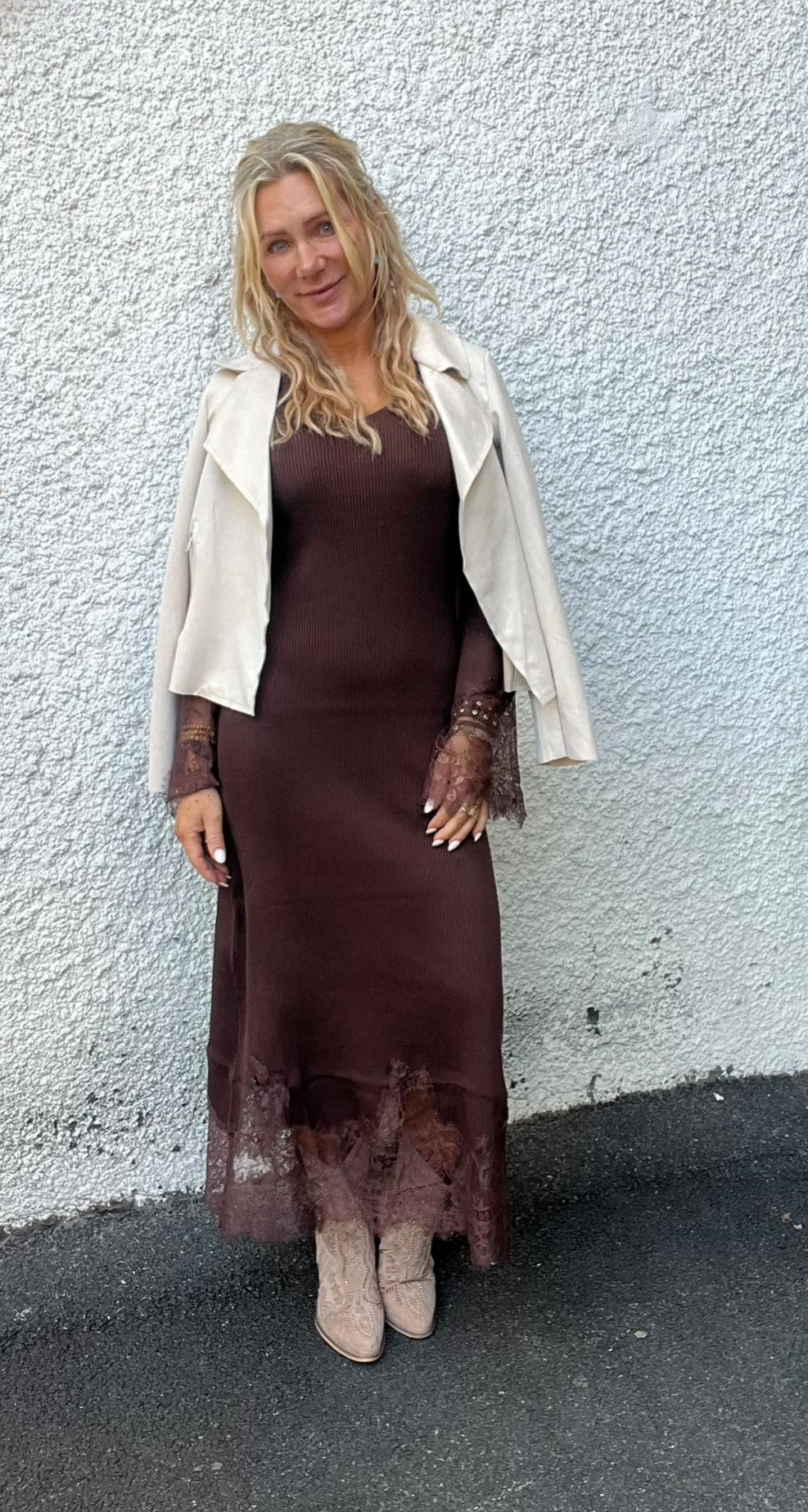 Kvinna med långt blont hår står och poserar mot en ljus, grovputsad vägg. Hon bär en lång brun klänning med spetsdetaljer, ljus jacka över axlarna och beige boots, och ler mot kameran.