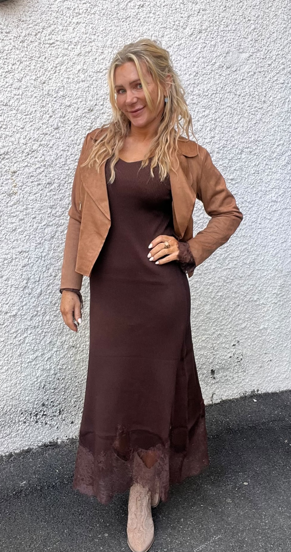 Leende kvinna poserar med handen på höften framför en ljus, grov putsad vägg. Hon bär en lång brun klänning med spetskant, kort brun jacka och ljusa boots, med blont vågigt hår.