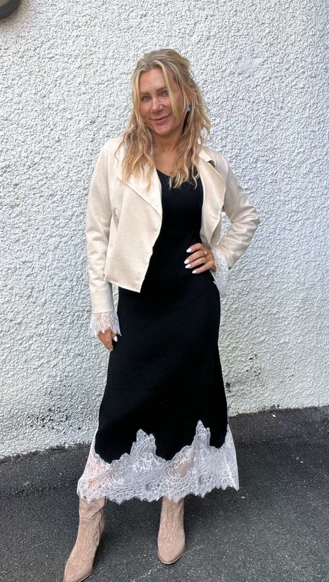 Kvinna poserar leende med handen på höften framför en ljus, grovputsad vägg. Hon bär svart ribbad klänning med vit spetskant nertill, ljus kort jacka med spetsmuddar och beige boots.