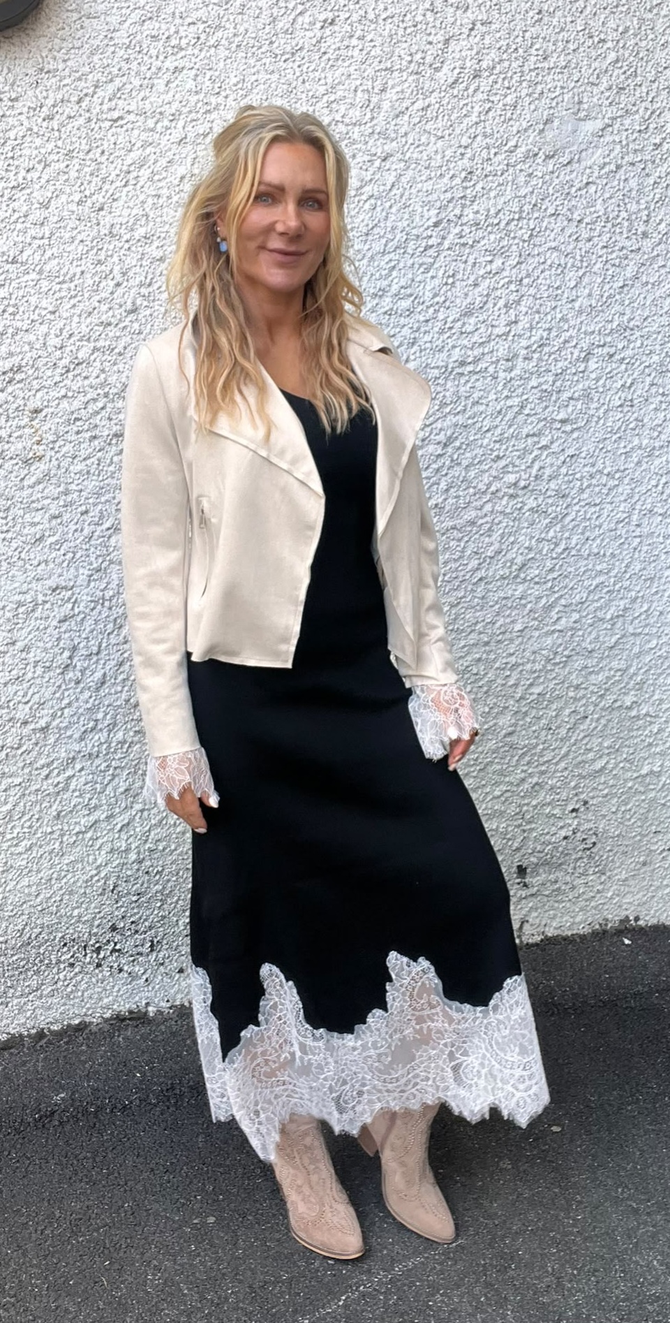 Kvinna med långt blont hår står och ler mot kameran framför en ljus, grovputsad vägg. Hon bär svart klänning med vit spetskant och manschetter, ljus kort jacka samt beige ankelboots.