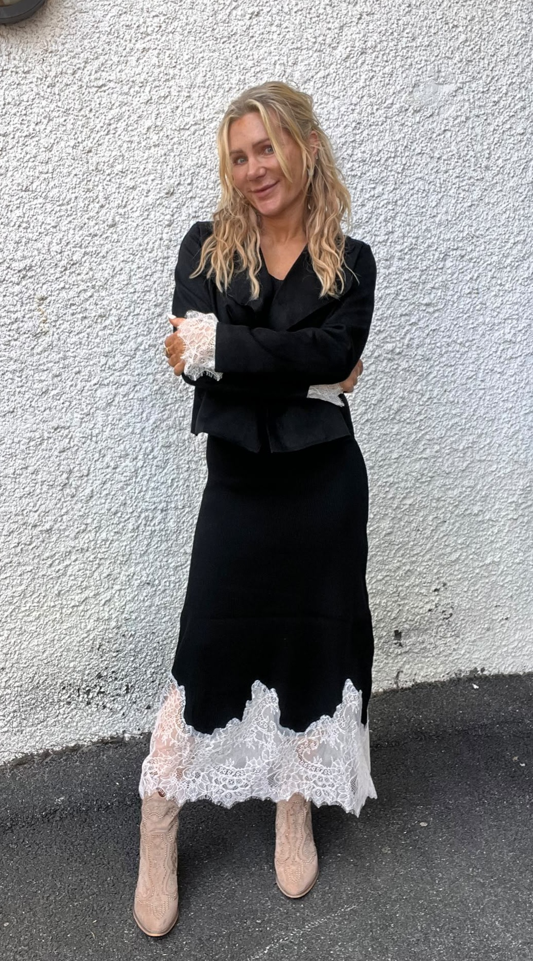 Kvinna med långt blont, vågigt hår står med armarna korsade och ler mot kameran framför en ljus, grovputsad vägg. Hon bär svart outfit med vita spetsdetaljer och beige cowboyboots.