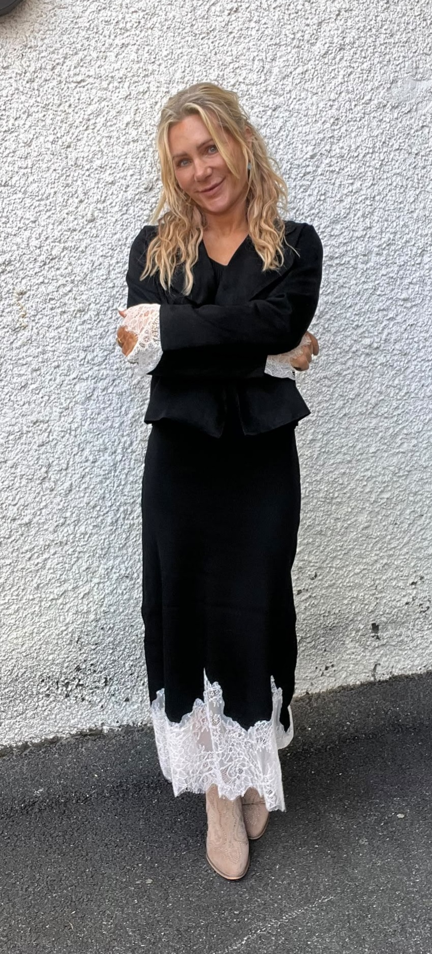 Kvinna med långt blont hår står med armarna i kors och ler lätt mot kameran. Hon bär svart klänning med vit spets vid ärmar och nederkant samt ljusa boots, framför en vit putsad vägg utomhus.