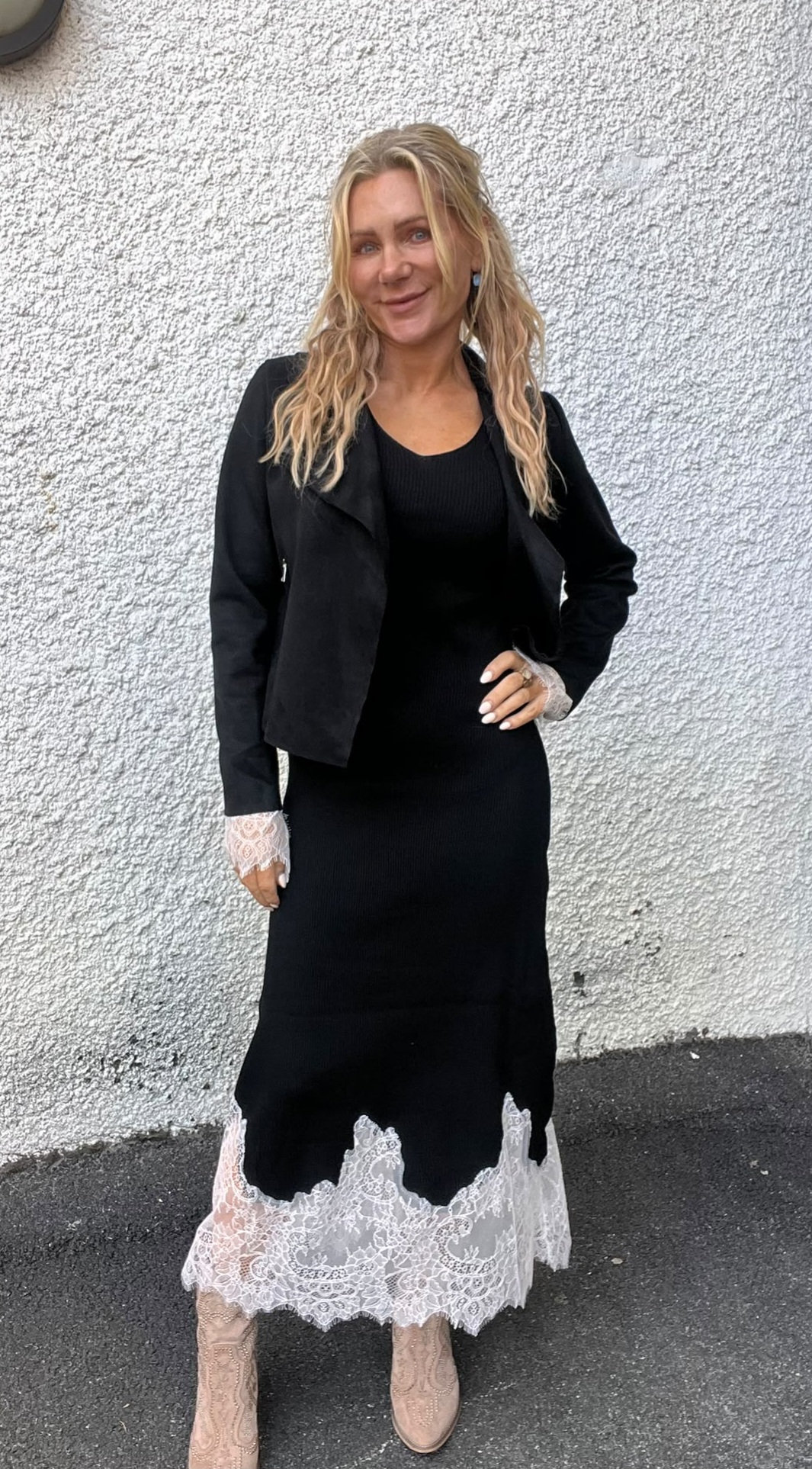 Kvinna med långt blont hår poserar och ler mot kameran med ena handen på höften. Hon bär svart klänning med vit spetskant och svart jacka, beige boots, och står mot en ljusputsad vägg utomhus.