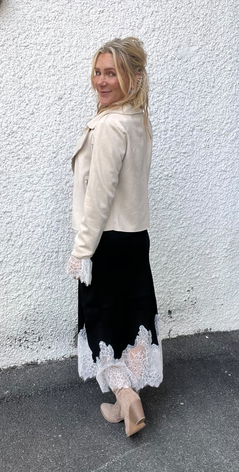 Blond kvinna står vänd i profil och tittar leende över axeln mot kameran, framför en vitputsad vägg. Hon bär ljus jacka, svart lång klänning med vit spetskant och ljusa boots.