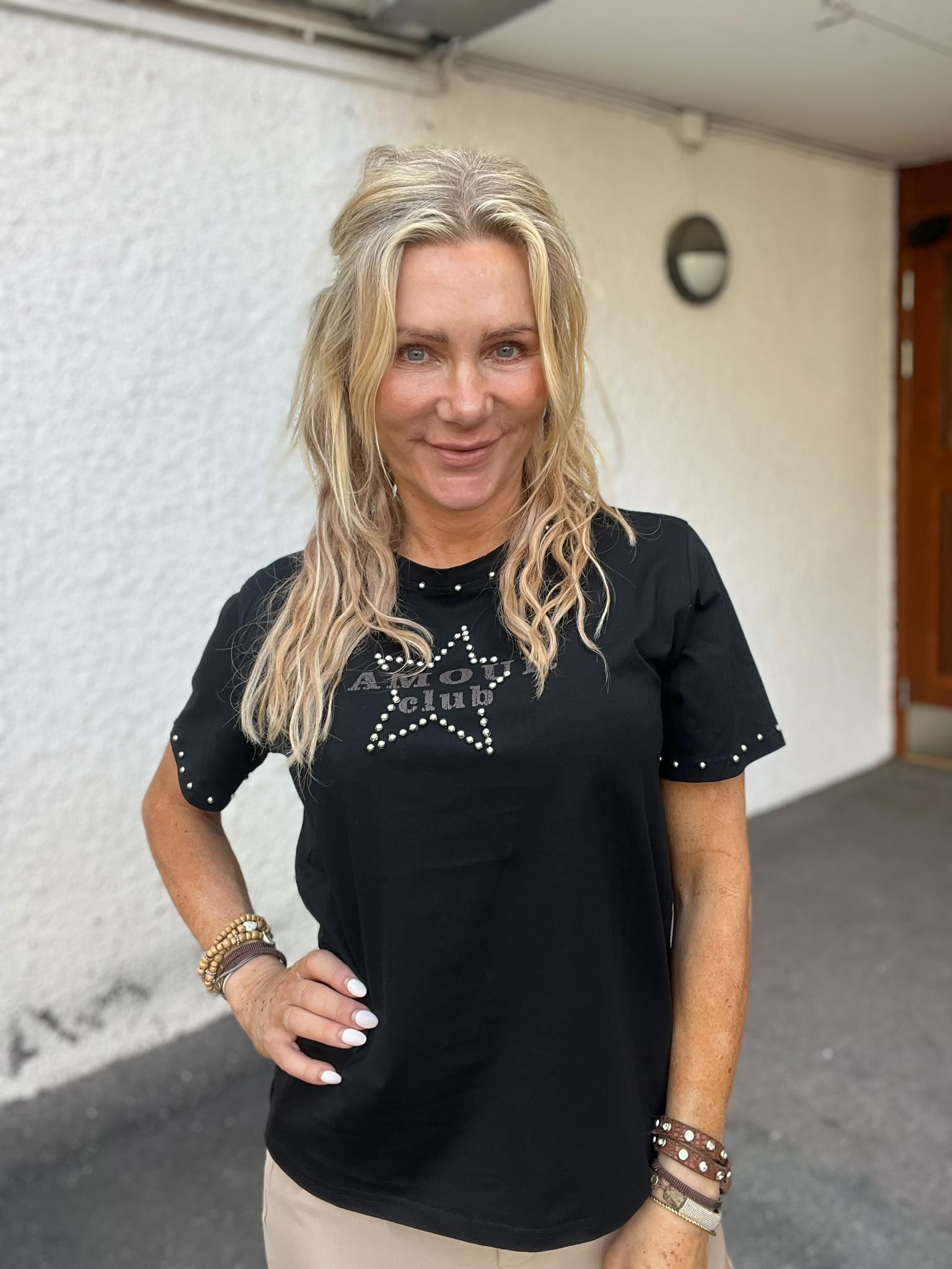 Leende blond kvinna poserar med handen på höften utomhus mot en ljus vägg och en dörr. Hon bär svart T-shirt med texten “AMOUR club” och en stjärna av nitar samt flera armband.