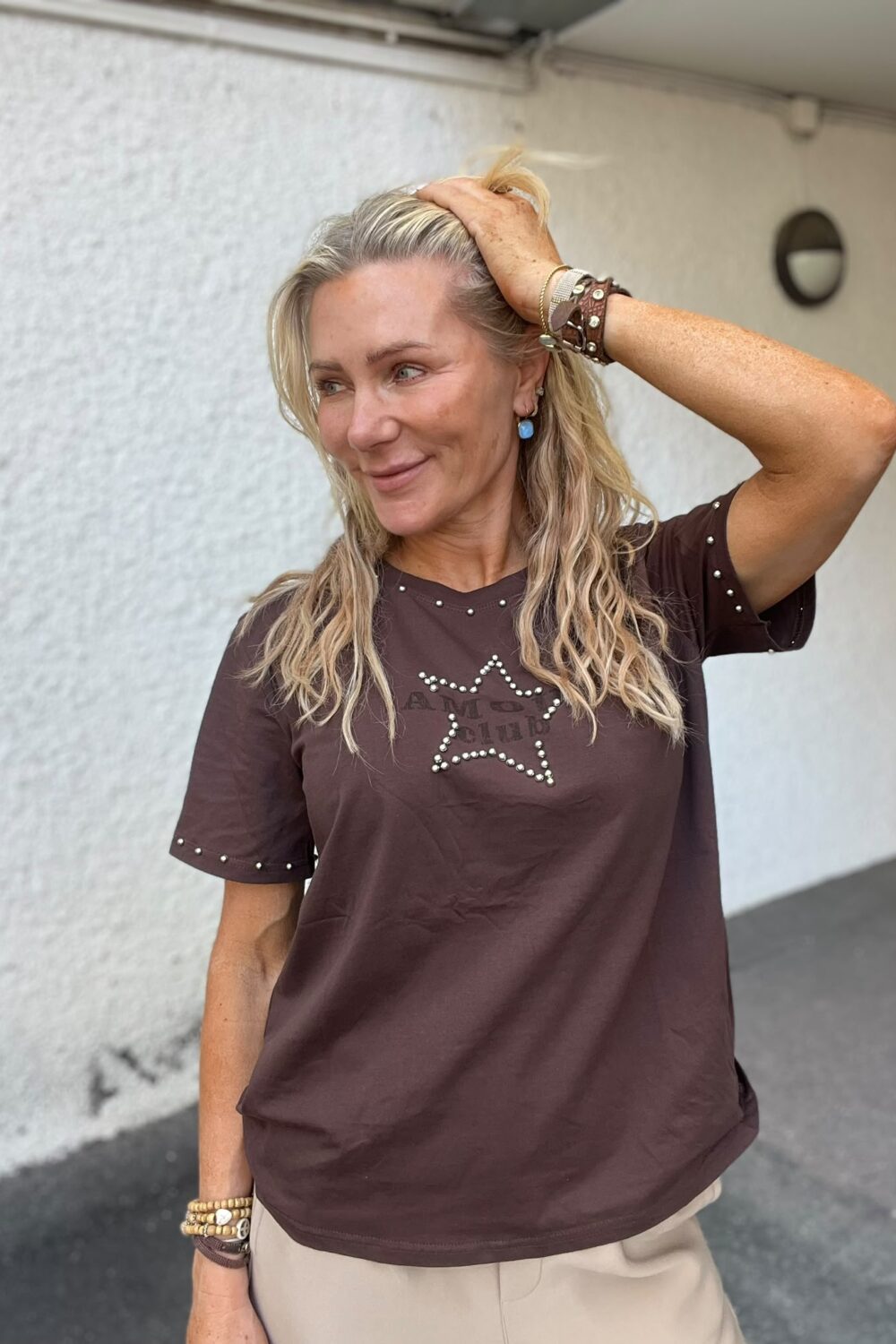 Blond kvinna står utomhus vid en ljus vägg, ler och håller ena handen i håret medan hon tittar åt sidan. Hon bär brun T-shirt med en nitarformad stjärna och texten “AMO RIM”, samt armband och blå örhängen.