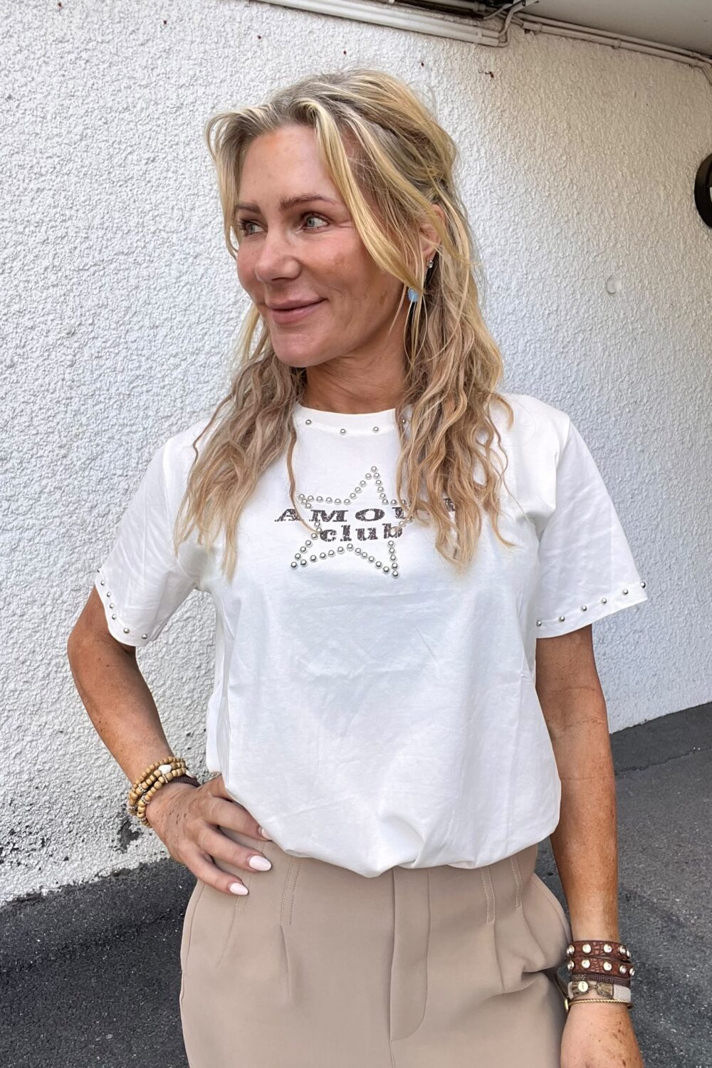 Leende kvinna med långt blont hår står med handen på höften och tittar åt sidan, framför en vit grovputsad vägg med en rund vägglampa. Hon bär vit T‑shirt med texten “AMOUR” och “CHIC”.
