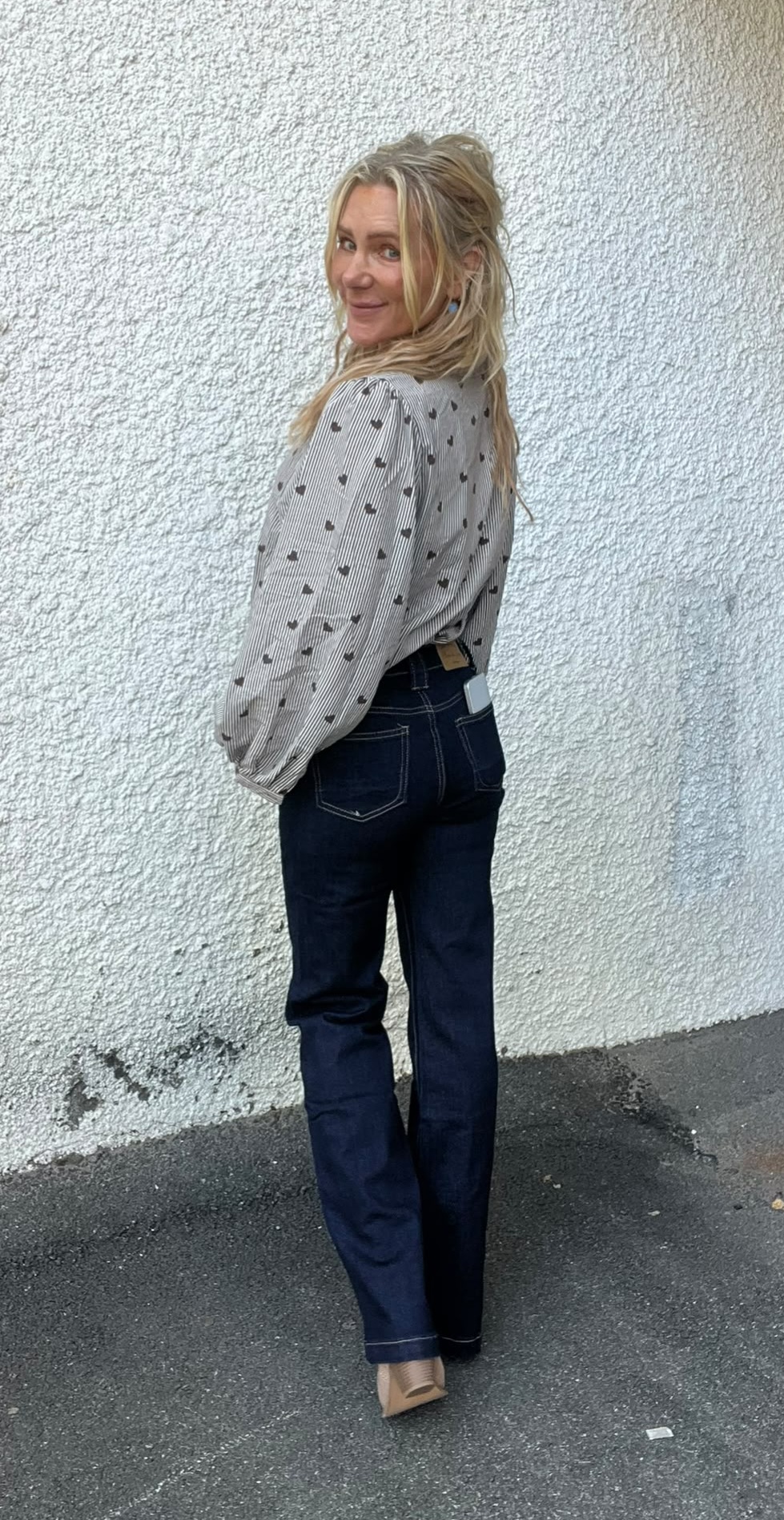Kvinna står utomhus med ryggen mot kameran och vänder huvudet för att le mot fotografen. Hon bär ljus prickig blus, mörka jeans och beige klackar, med mobil i bakfickan, framför vit putsad vägg.