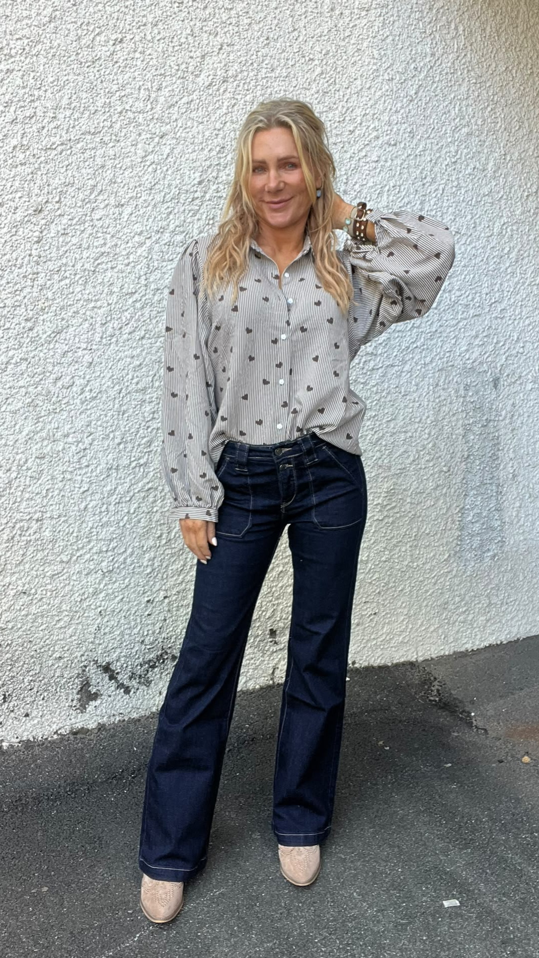 Kvinna med långt blont hår står mot en vit, grovputsad vägg och ler. Hon har ena handen bakom huvudet och bär randig blus med små hjärtan, mörkblå jeans och ljusa boots på asfalt.