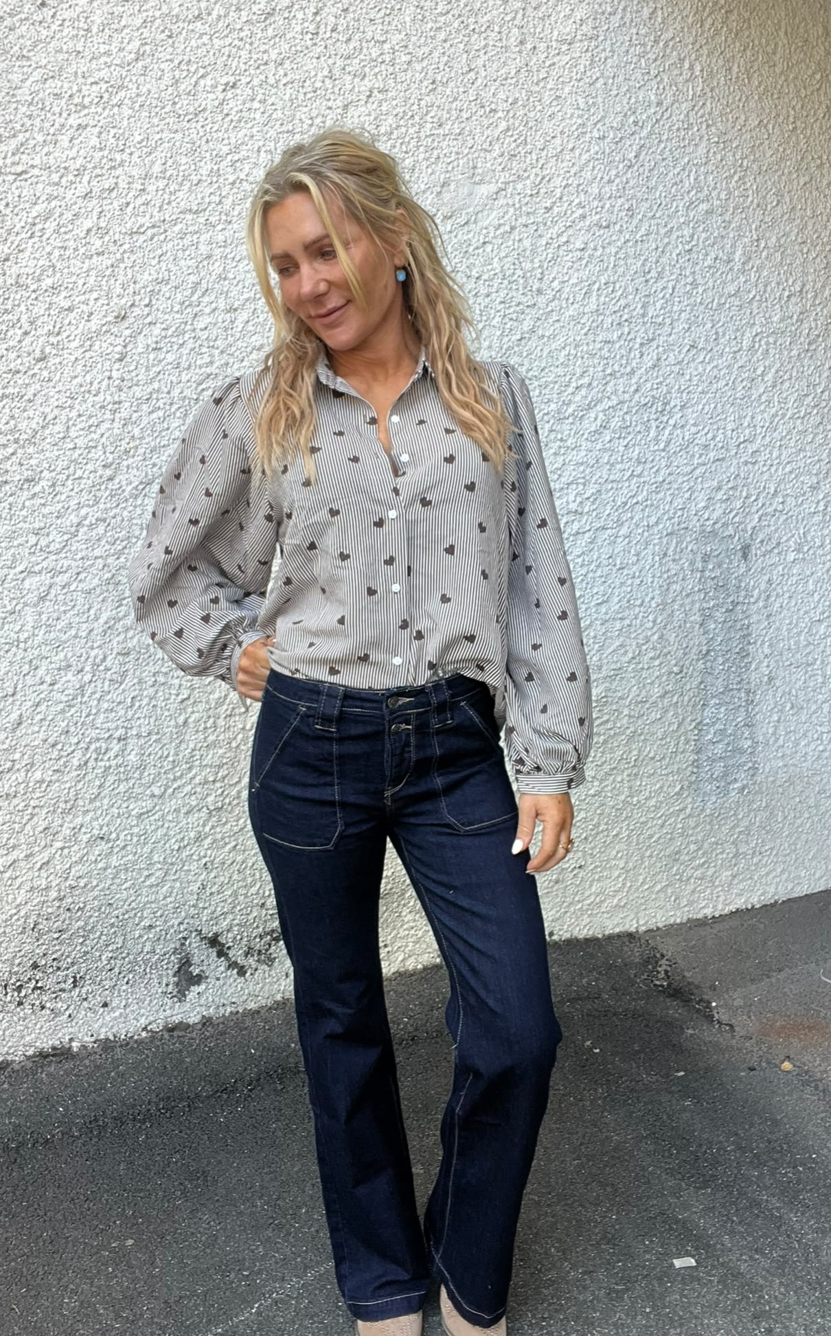 Kvinna med långt blont hår står avslappnat mot en ljus, grovputsad vägg och ser ned åt sidan. Hon har handen i fickan och bär randig skjorta med små hjärtan samt mörkblå jeans.