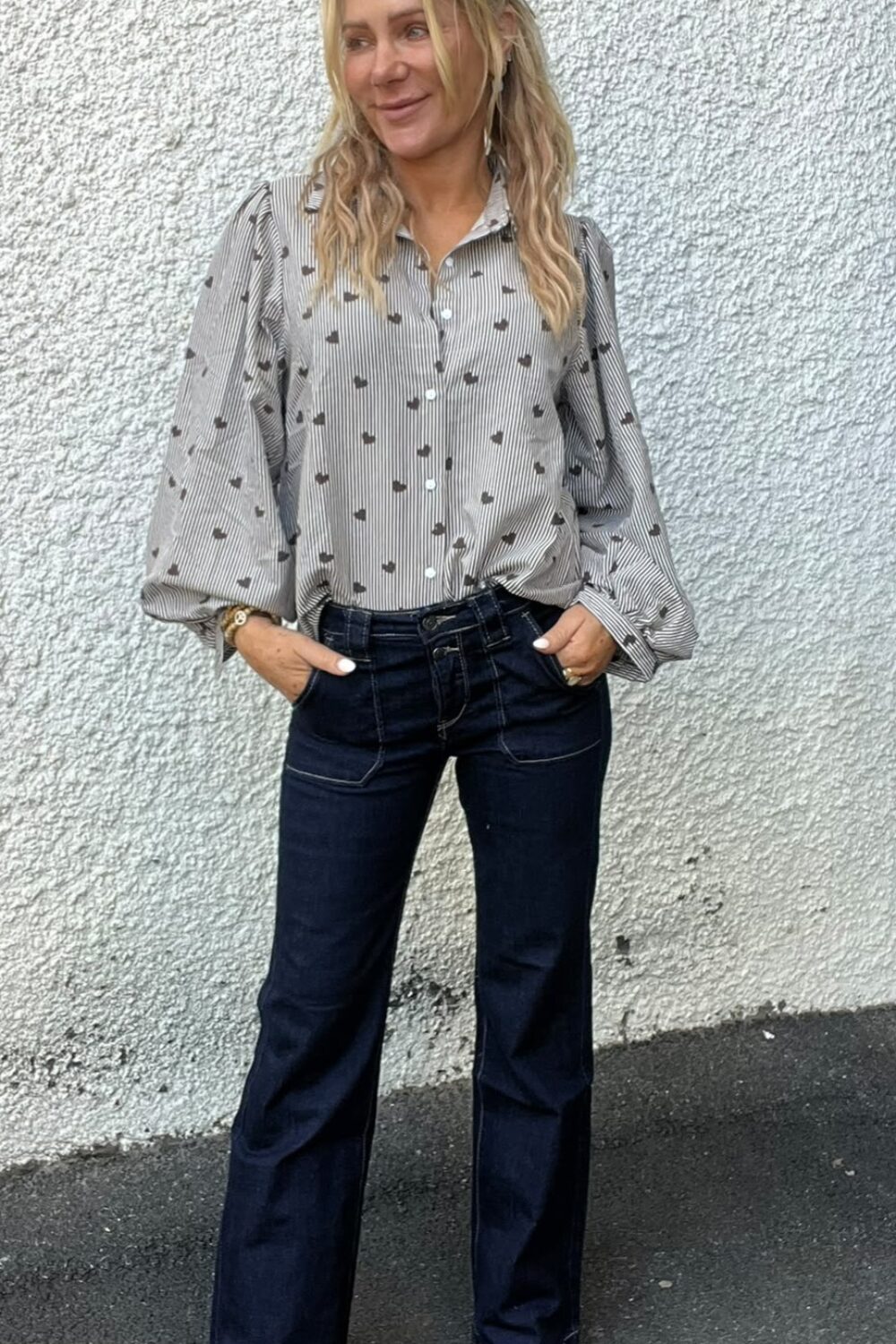 Kvinna med långt blont hår står avslappnat med händerna i fickorna och blickar åt sidan. Hon bär gråmönstrad skjorta med puffärmar, mörkblå utsvängda jeans och beige boots mot en grov vit vägg.