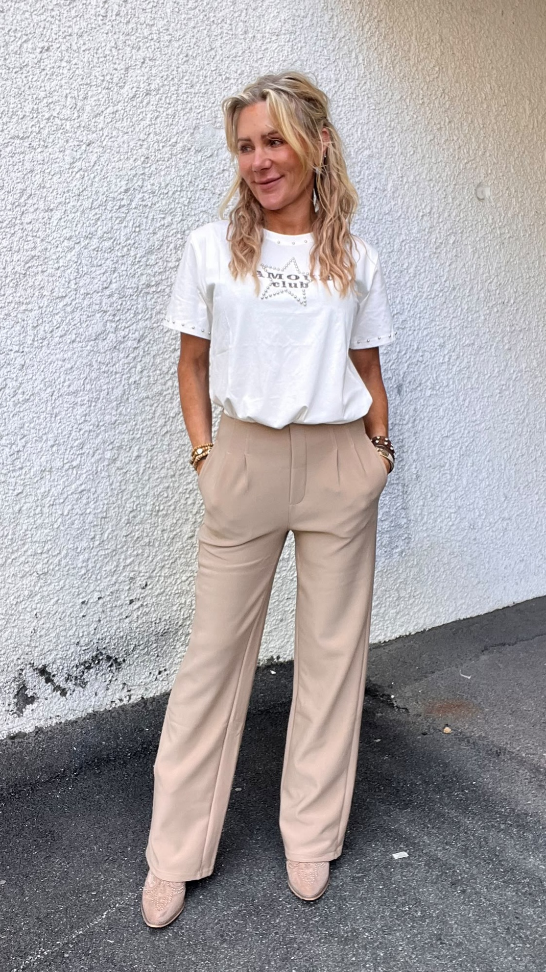 Blond kvinna står med händerna i fickorna och ler åt sidan framför en vit, putsad vägg. Hon bär vit T‑shirt med texten “M… club” i en stjärna, beige byxor och ljusa skor.