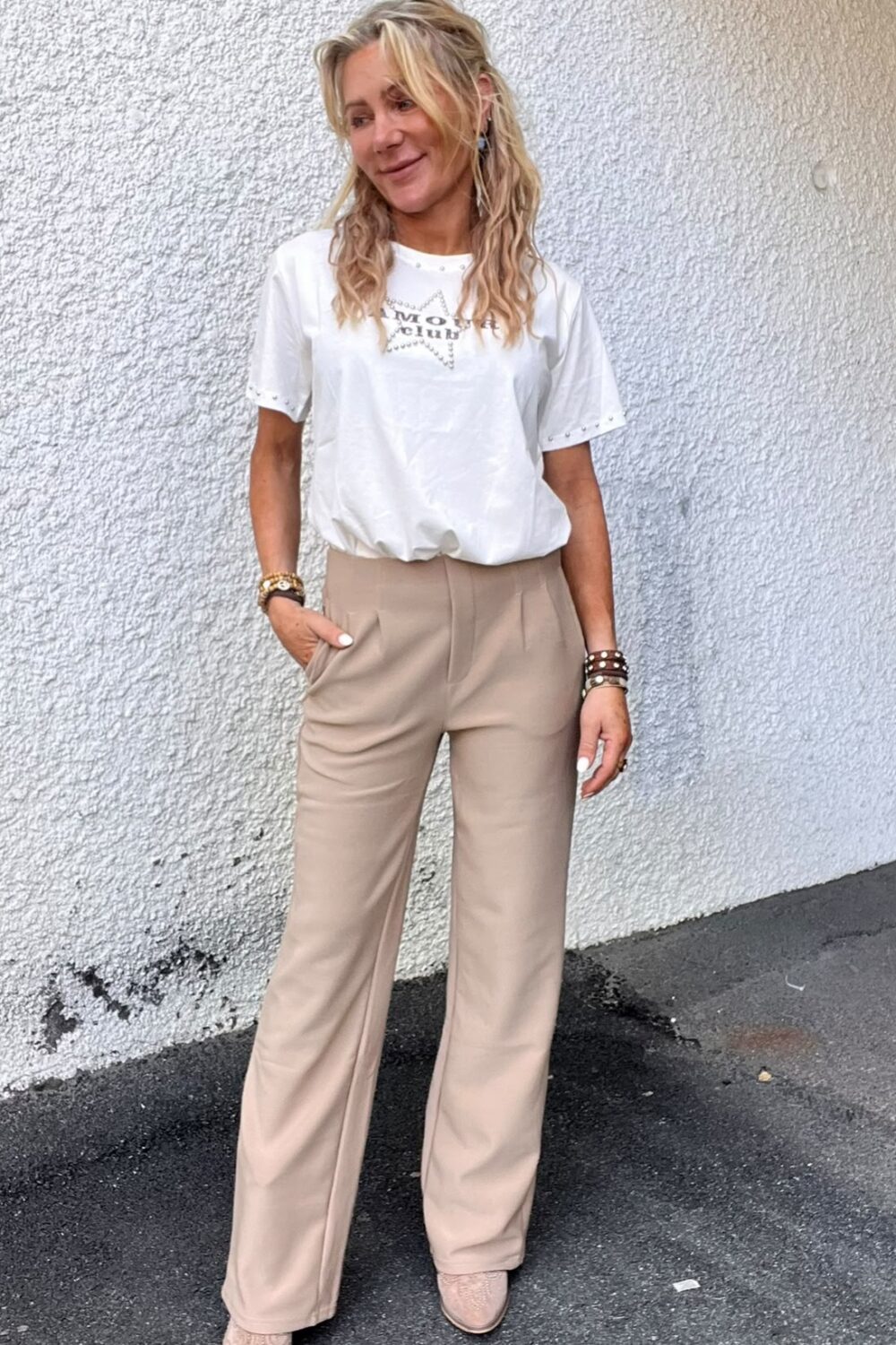 Kvinna med långt blont hår står avslappnat mot en ljus, grovputsad vägg och ler åt sidan, med ena handen i fickan. Hon bär vit t-shirt med texten ”AMOUR” och beige byxor samt armband.