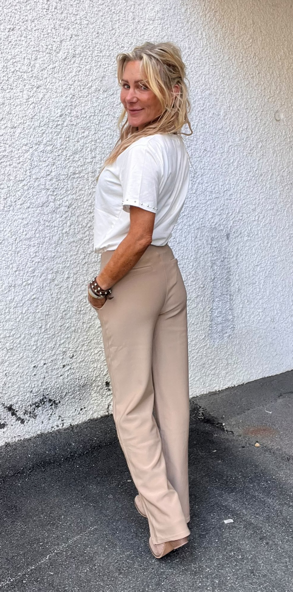 Blond kvinna står i profil med händerna i fickorna och ler mot kameran. Hon bär vit kortärmad topp med små nitar och beige byxor, framför en vit, grovputsad vägg på asfalt utomhus.