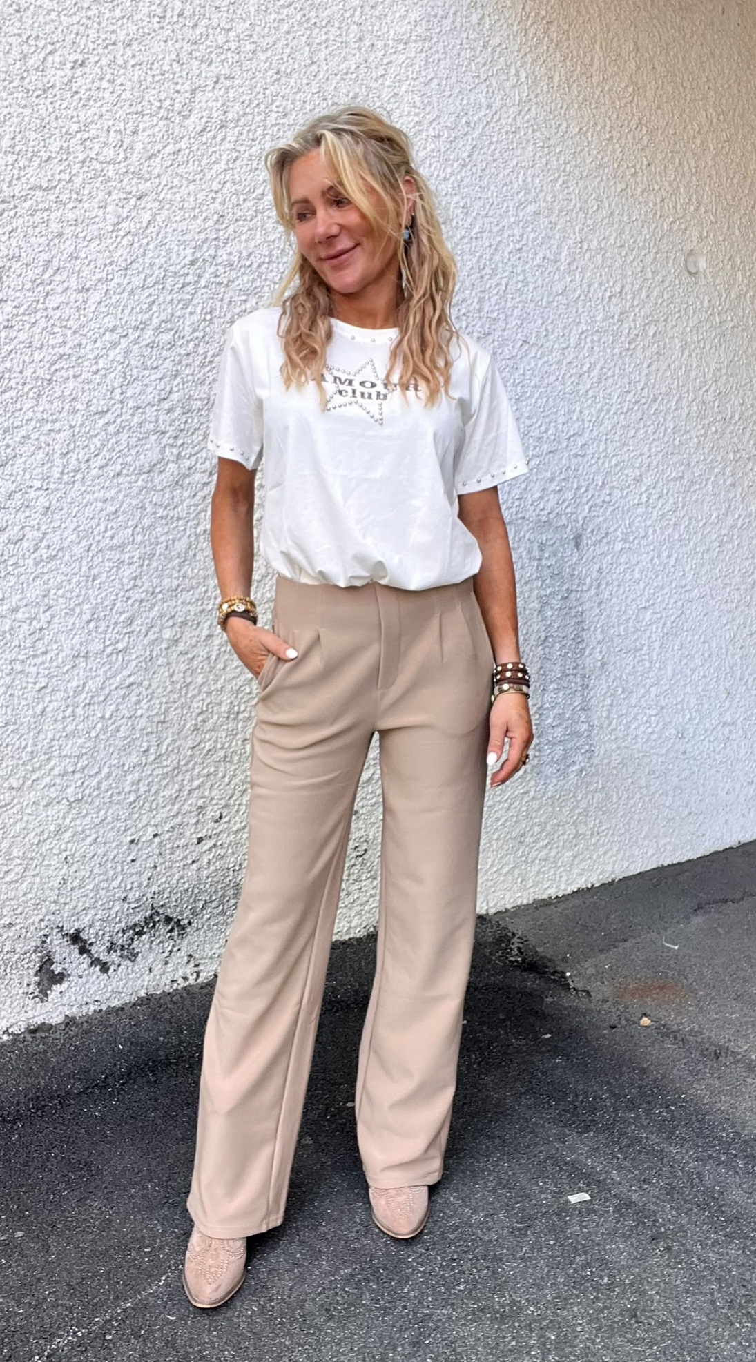 Kvinna med långt blont hår står avslappnat mot en ljus, grovputsad vägg och ler åt sidan, med ena handen i fickan. Hon bär vit t-shirt med texten ”AMOUR” och beige byxor samt armband.