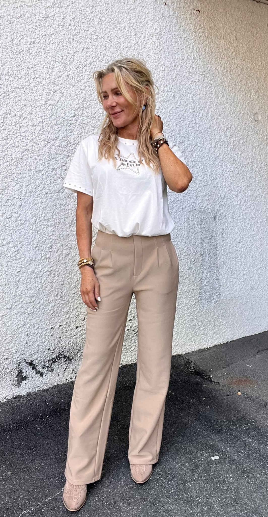 Blond kvinna står avslappnat mot en vit, grovputsad vägg, ler och rör vid håret. Hon bär vit T-shirt med texten “WAY OF LIFE”, beige byxor, armband och ljusa skor, på asfalt utomhus.