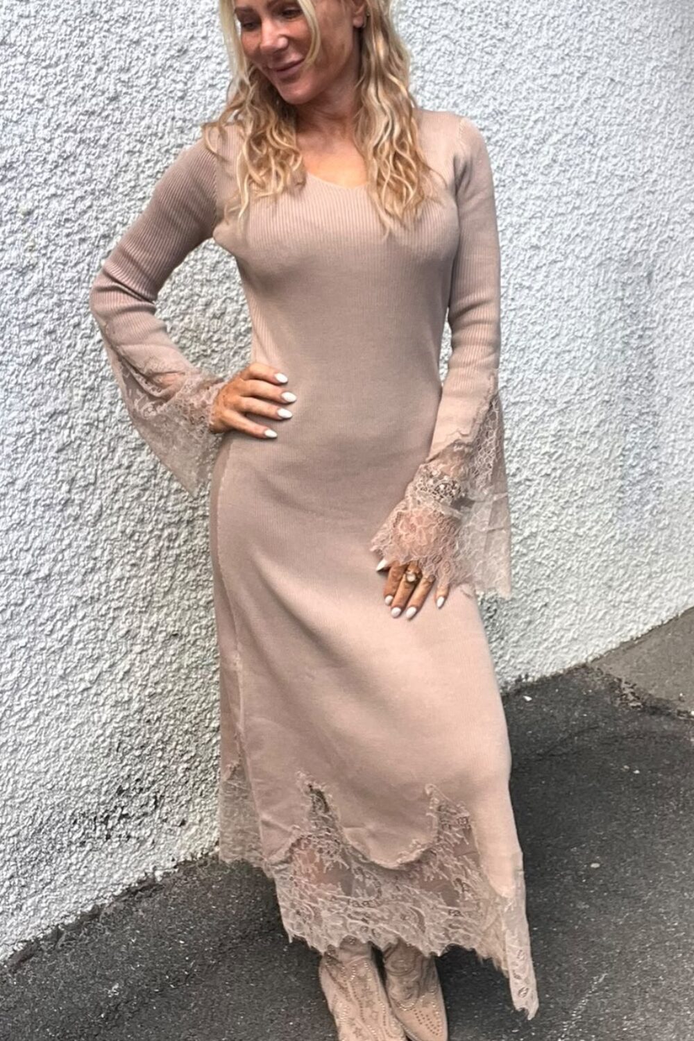 Leende kvinna poserar med ena handen på höften vid en grov vit vägg. Hon bär en lång, beige stickad klänning med spetsdetaljer vid ärmar och nederkant samt matchande ljusa boots på asfalt.