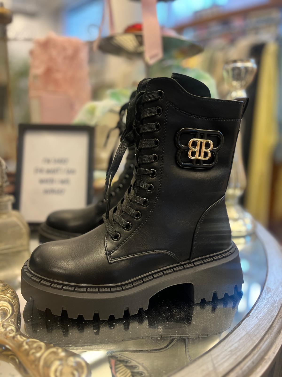 Svart snörkänga i läder står uppställd på en glasbricka i en butik. Den har grov, tjock sula med kraftigt mönster och en guldfärgad “BB”-logotyp på utsidan; bakgrunden är suddig med andra föremål.