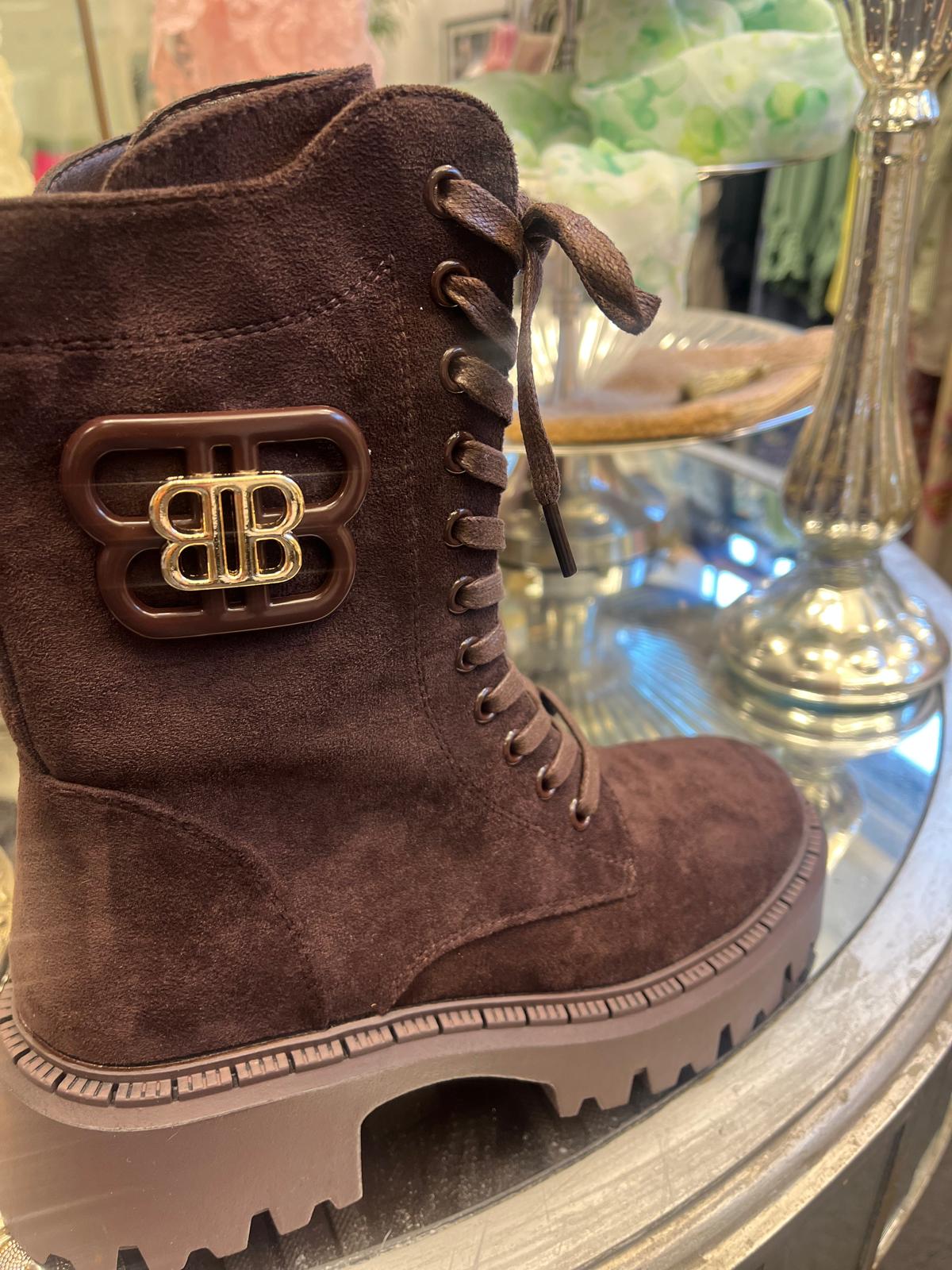 Brun snörkänga i mocka står uppställd på en glasskiva, med grov räfflad sula och metallöljetter. På sidan sitter ett stort spänne med guldfärgad logotyp “BB”, i en butikslik miljö.
