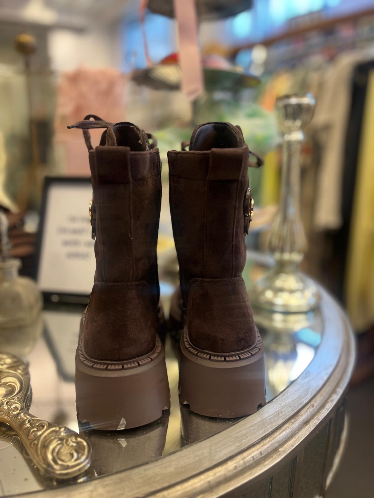 Ett par mörkbruna ankelboots i mocka står bredvid varandra med baksidan mot kameran på ett runt glasbord. De har tjocka plattformsulor och dragflik baktill, i en butiksmiljö med suddig inredning.