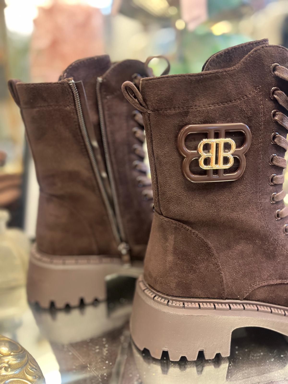 Två bruna mockakängor står uppställda i en butiksmiljö; den främre visar snörning och en metalllogga med texten “BB”, medan den bakre har en synlig dragkedja. Kraftiga, ljusbruna sulor och suddig bakgrund.