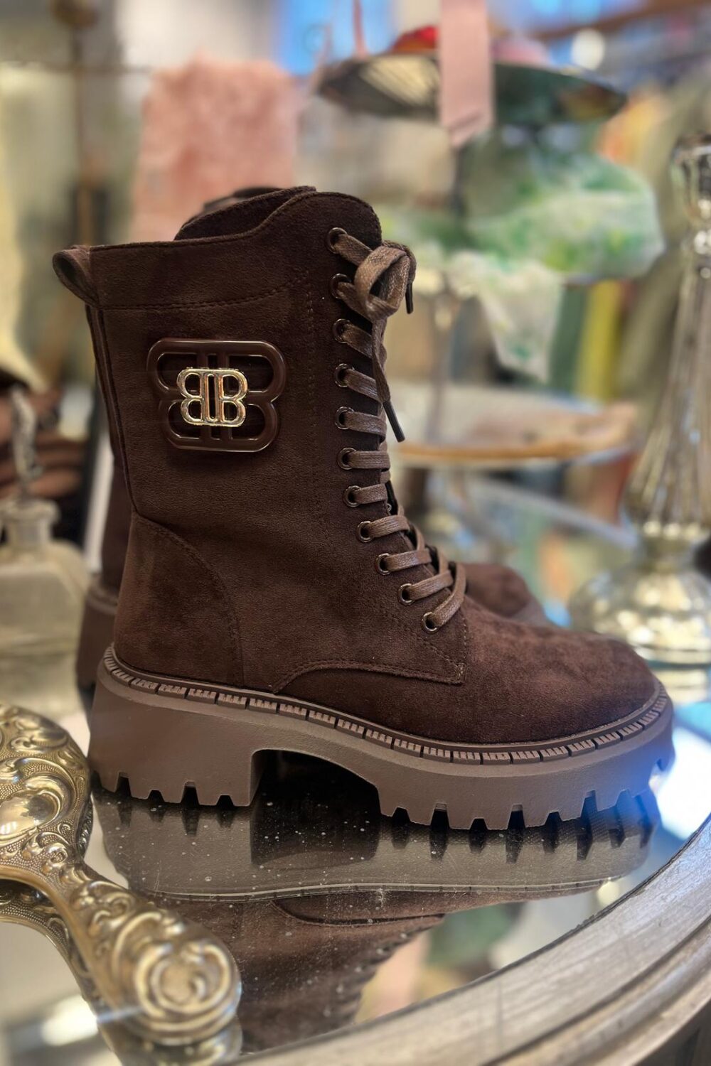 Brun snörkänga i mocka står uppställd på ett runt glassbord och visas i profil, med kraftig räfflad platåsula och metallmärke med bokstäverna “BB” på skaftet; butiksmiljö i mjuk oskärpa.
